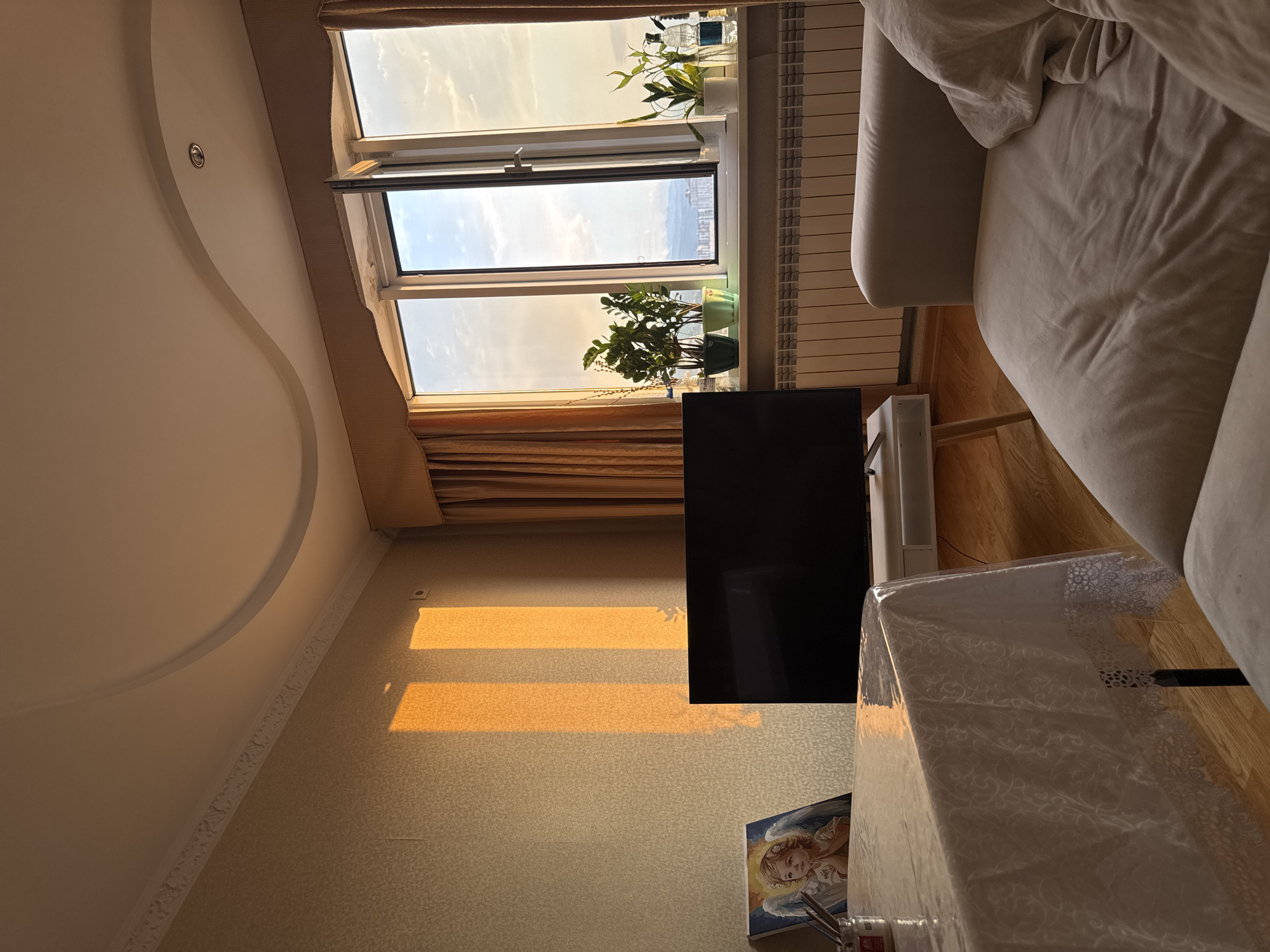 The image depicts an interior scene, likely a living room, bathed in the warm glow of sunset. Sunlight streams through an open window, casting elongated rectangular shadows on a textured, light-colored wall. The window overlooks a city skyline with distant mountains, suggesting an urban environment. Inside, a light-colored sofa occupies the foreground on the right. To the left, a dining table is covered with a translucent white tablecloth with a lace trim. On the table, an artwork depicting an angel is visible, along with a small jar and possibly some art supplies. A black flat-screen television is placed on a white media console in the center of the room. On the windowsill, several potted plants and bottles are arranged. The ceiling features a decorative, curved design. The overall impression is one of a tranquil evening in a well-lit apartment. Without specific landmarks or context, pinpointing the exact location in Almaty, Kazakhstan is not possible from this image alone.