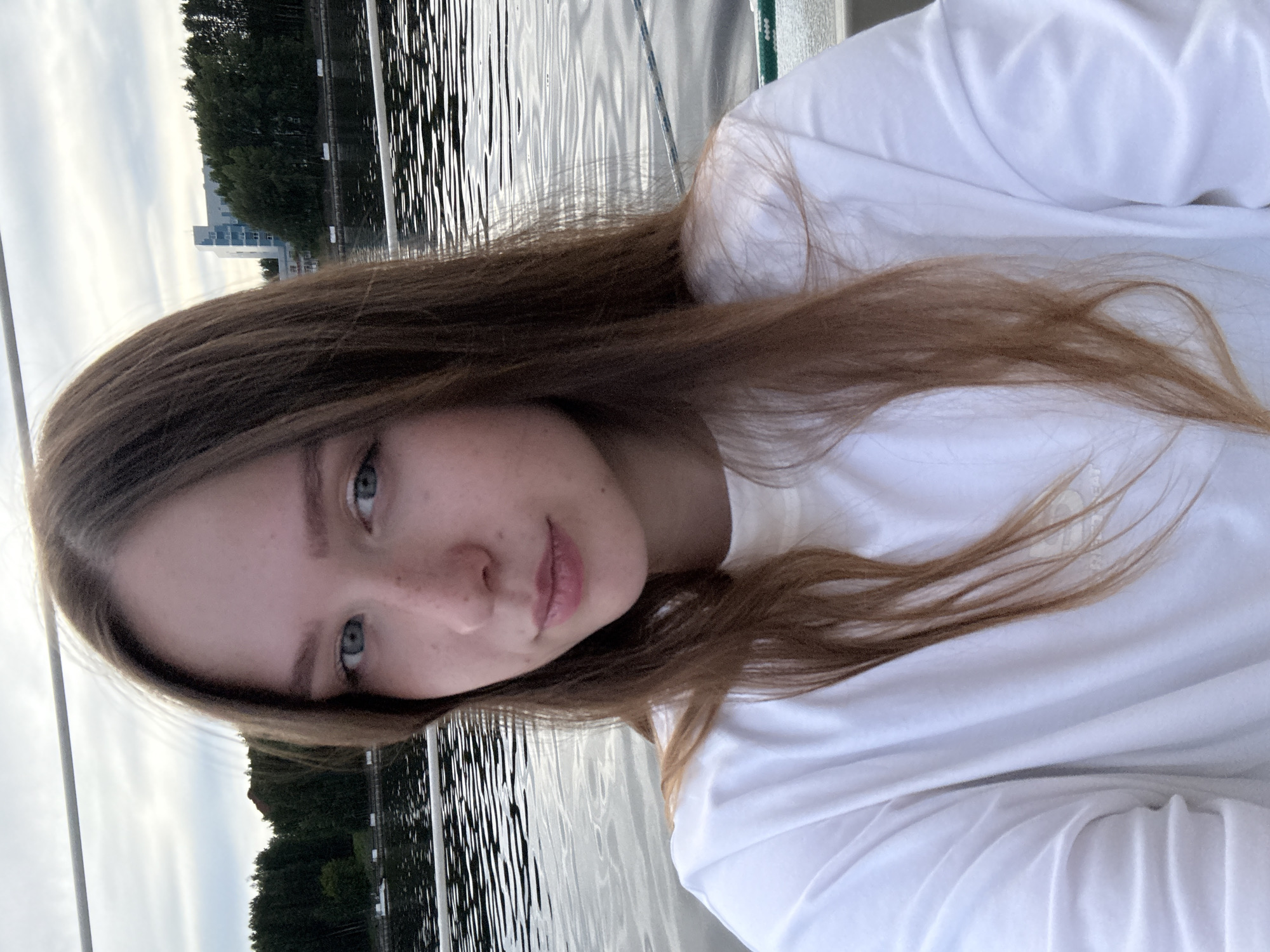 A young woman with long, light brown hair and blue eyes is captured in a close-up selfie. She is wearing a white t-shirt. The background consists of rippling water, a tree-lined shore, and a modern building. The setting appears to be a body of water, possibly a river or lake, near an urban area. The overall impression is a casual outdoor portrait. This scene is located on the Kama River, within the city of Chaikovsky, Russia.