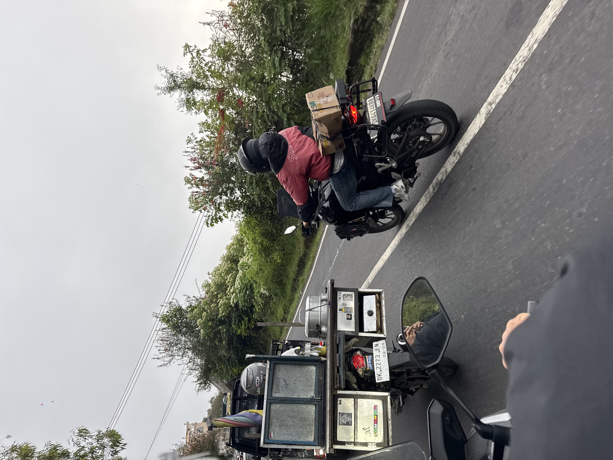 A motorcycle carrying two people is traveling down a paved road in Kintamani, Indonesia. The rider is wearing a red jacket and blue jeans, and the passenger is wearing a black jacket and helmet. The motorcycle is equipped with a rack that holds two cardboard boxes. In the foreground, another motorcycle's handlebar and mirror are visible, reflecting a person's hands holding the handlebar and a green, leafy background. To the left of the road, a modified pickup truck with a food cart attached is parked. The food cart has a silver-colored exterior with various containers and equipment. A rainbow-colored flag is attached to the truck. The environment is lush and green, with trees and vegetation lining the road. The sky is overcast and appears to be raining lightly, as evidenced by the wet road surface and the overcast conditions.