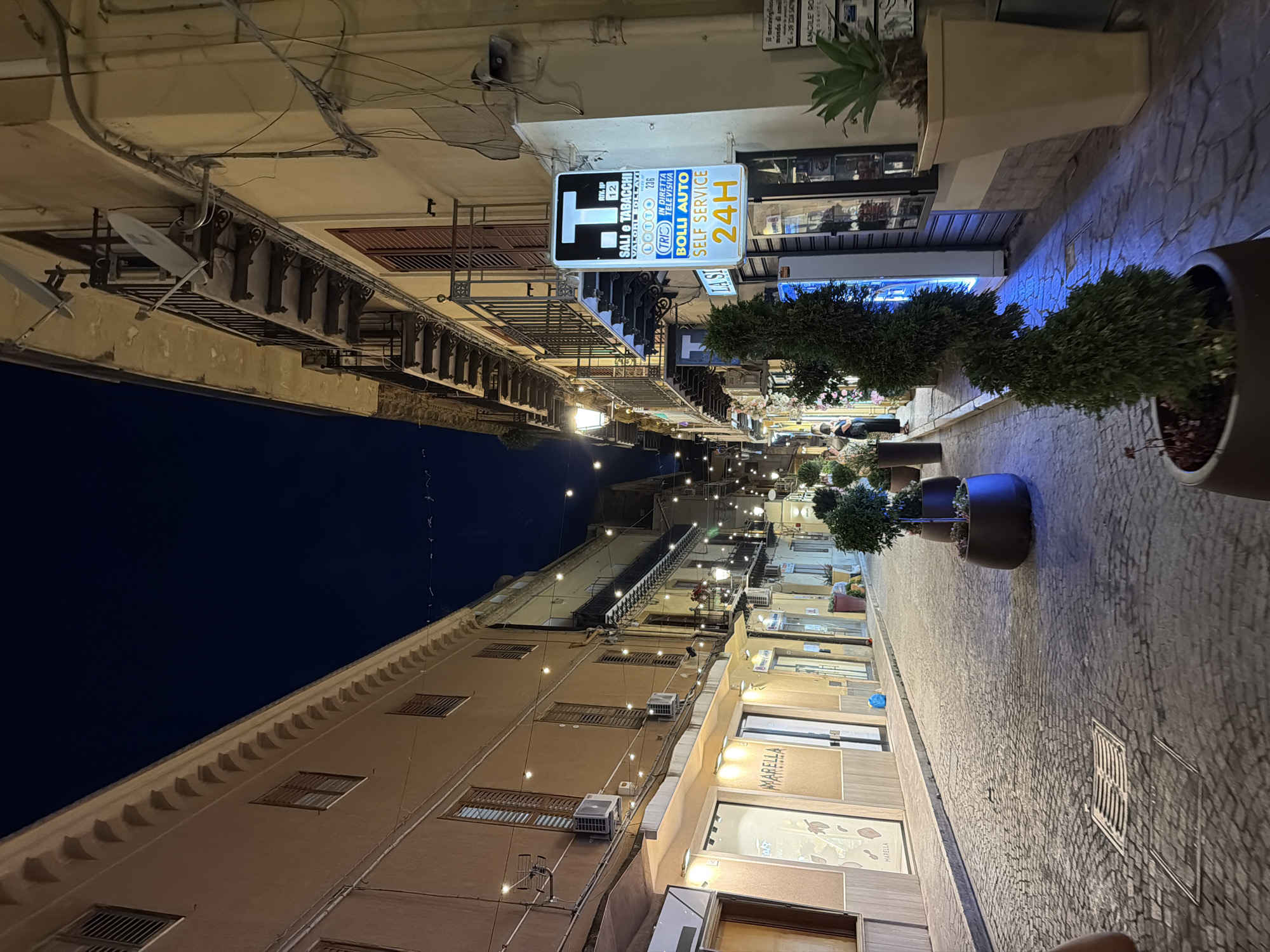 The image depicts a narrow cobblestone street in Agrigento, Italy, at night. Buildings with shuttered windows and balconies line both sides of the street, illuminated by string lights and the warm glow from shopfronts. One prominent sign on the right advertises "SALI e TABACCHI" (tobacco and salt shop) along with lottery information and a "SELF SERVICE 24H" service. Several businesses, including "MARELLA," are visible. Potted plants, including a spiral-shaped evergreen, are placed along the street and in front of shops, adding a touch of greenery to the urban landscape. A few people are visible walking on the sidewalk, with one person in the foreground partially captured. The overall atmosphere is tranquil and inviting, typical of a European street scene after dark.