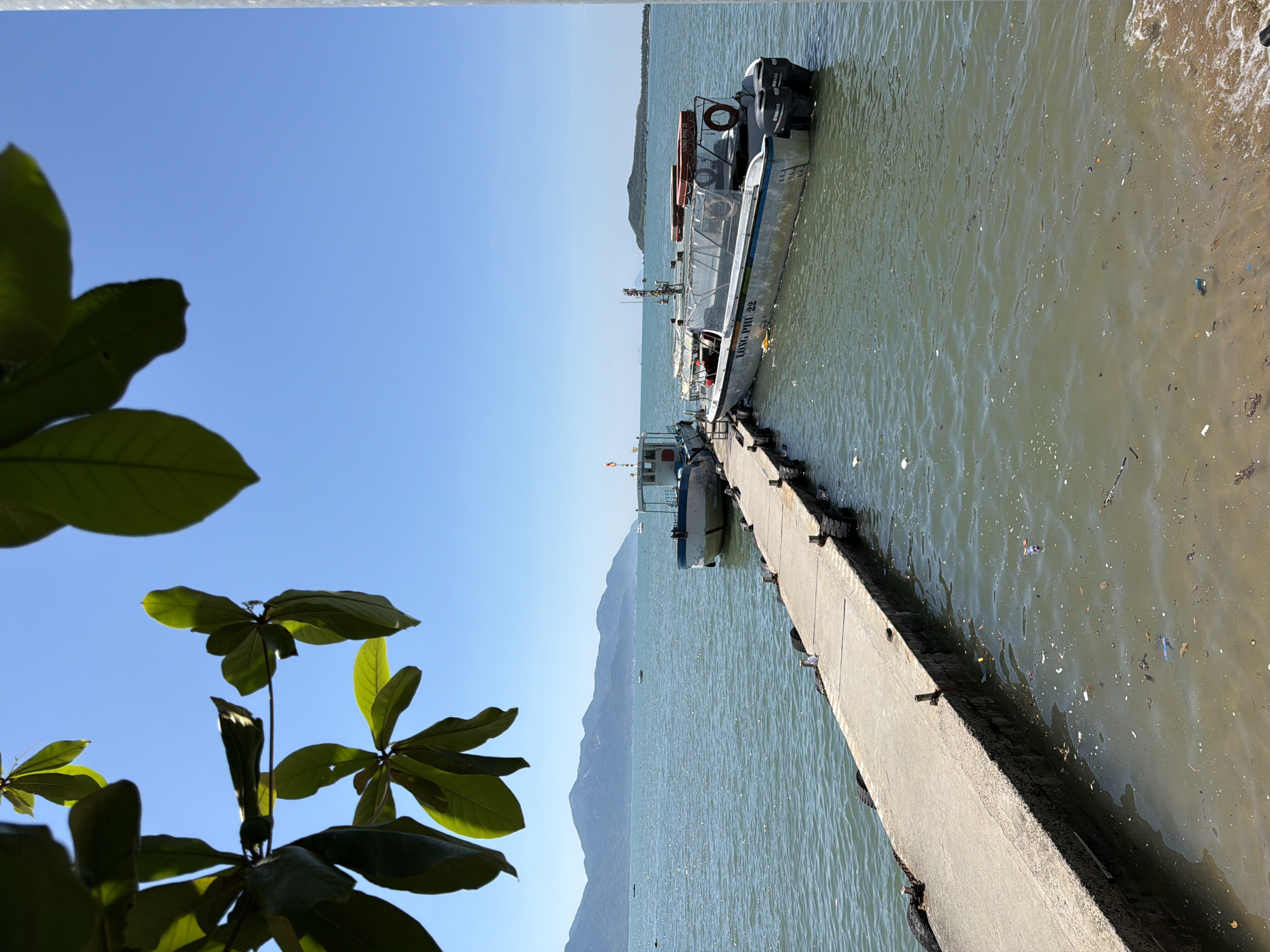 The image depicts a concrete pier extending into the ocean, with two boats moored to it. The pier is lined with black fenders and has several metal posts. The water around the pier is murky and contains visible debris, including plastic bottles and other refuse. In the background, mountains rise from the sea under a clear blue sky. The foreground features green leaves from a tree, framing the top left portion of the image. One of the boats, labeled "LONG PHU 22", is a long, white vessel with a covered seating area and two outboard motors at the stern. The other boat, to its left, is shorter and appears to be a different type of vessel. The scene suggests a coastal location, likely in the vicinity of Ninh Ích, Vietnam, given the context.