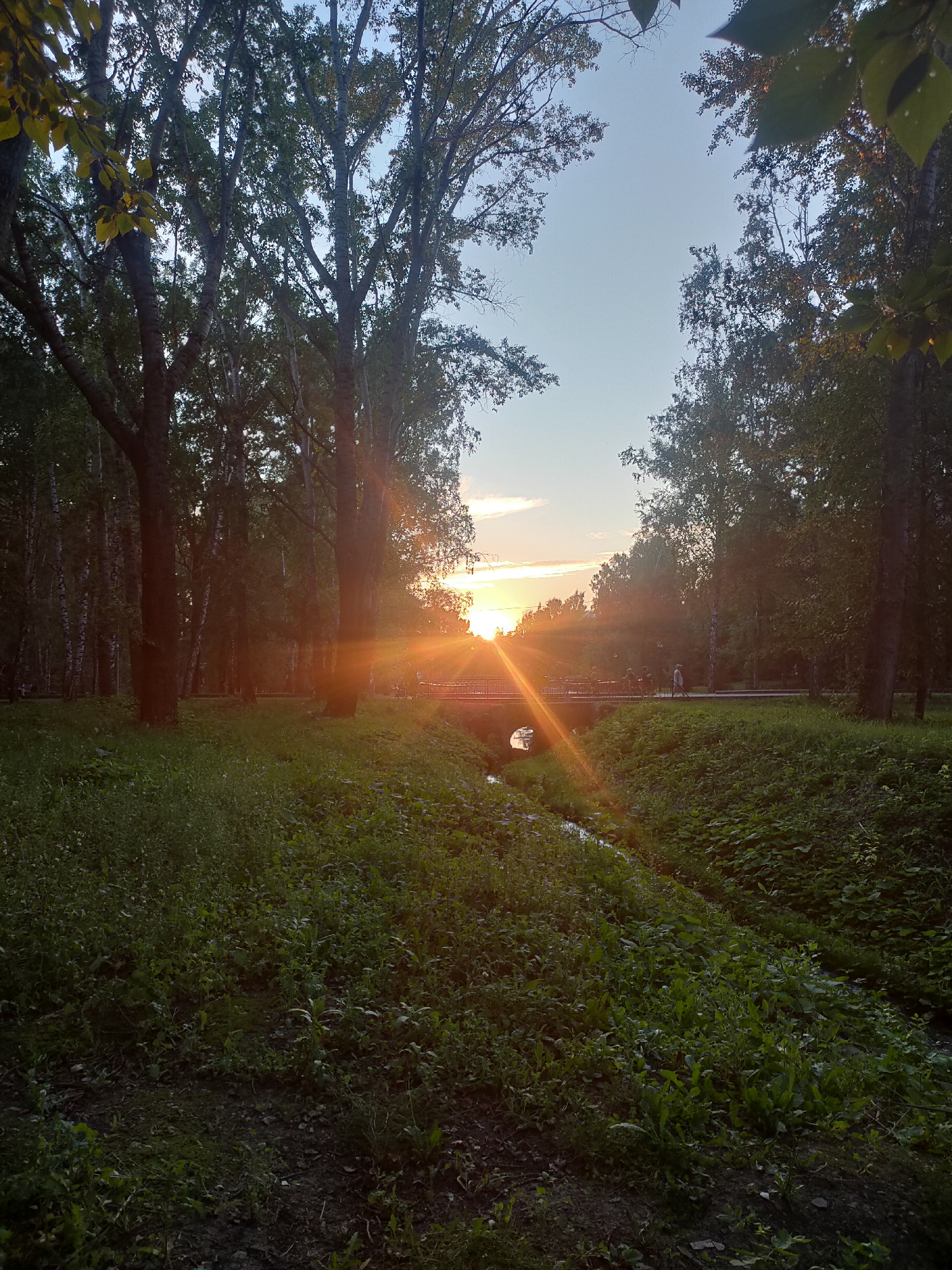 The image depicts a park scene in Oktyabrsky, Russia, during sunset. The sun, low on the horizon, casts a strong golden light with prominent sun flares, illuminating the sky and creating a lens flare effect across the scene. The foreground is dominated by lush green grass and various plants, with a narrow stream or ditch running through it. Trees with dense foliage line the park, framing the view. In the mid-ground, a small bridge spans the stream, and several individuals are visible walking or standing on or near the bridge, appearing as silhouettes against the bright sunset. The overall atmosphere is peaceful and natural, capturing a serene evening in a park.