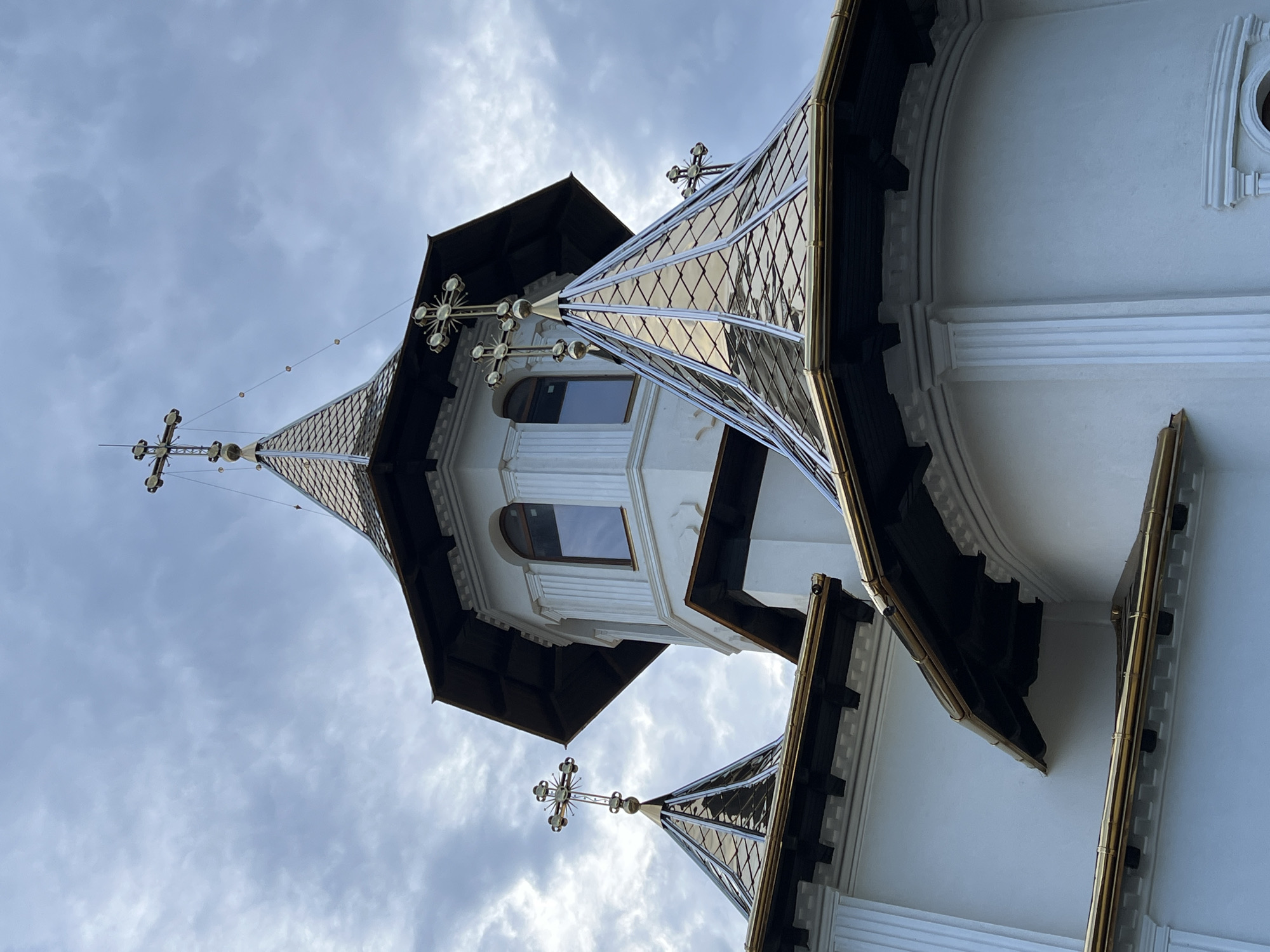 This image shows a low-angle, close-up view of the roofs and a tower of a white church, likely the Assumption of the Virgin Mary Church in Năvodari, Romania, against a cloudy sky. The church features multiple tiered roofs covered in golden, diamond-shaped tiles, accented with black trim and gold gutters. Several ornate crosses adorn the peaks of these roofs. A central tower rises above, also white, with arched windows and decorative fluting. The architectural style appears to be Eastern Orthodox. There are no people visible in the image. The environment is an overcast day, with a diffused light source.