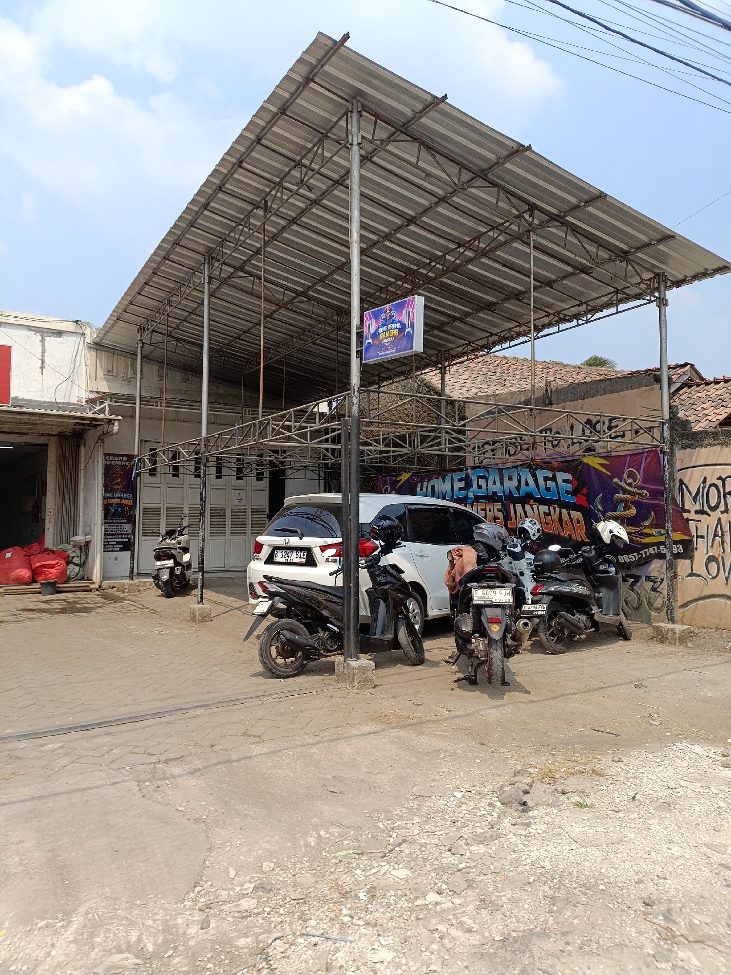 The image depicts an outdoor scene in what appears to be a commercial area, likely a garage or workshop, in Cariu, Indonesia. A metal-roofed structure with a supporting metal frame extends over a paved area. Several motorcycles and a white car are parked underneath this structure. To the left, a building with an open entrance is visible, with large red bags stacked inside. A sign is mounted on the wall of this building. On the right side of the image, a wall is adorned with graffiti, including large text that reads "HOME GARAGE" and "GAMERS JANGKAR," along with a phone number. The ground is a mixture of paved and unpaved surfaces, with some debris present. The sky above is partly cloudy.