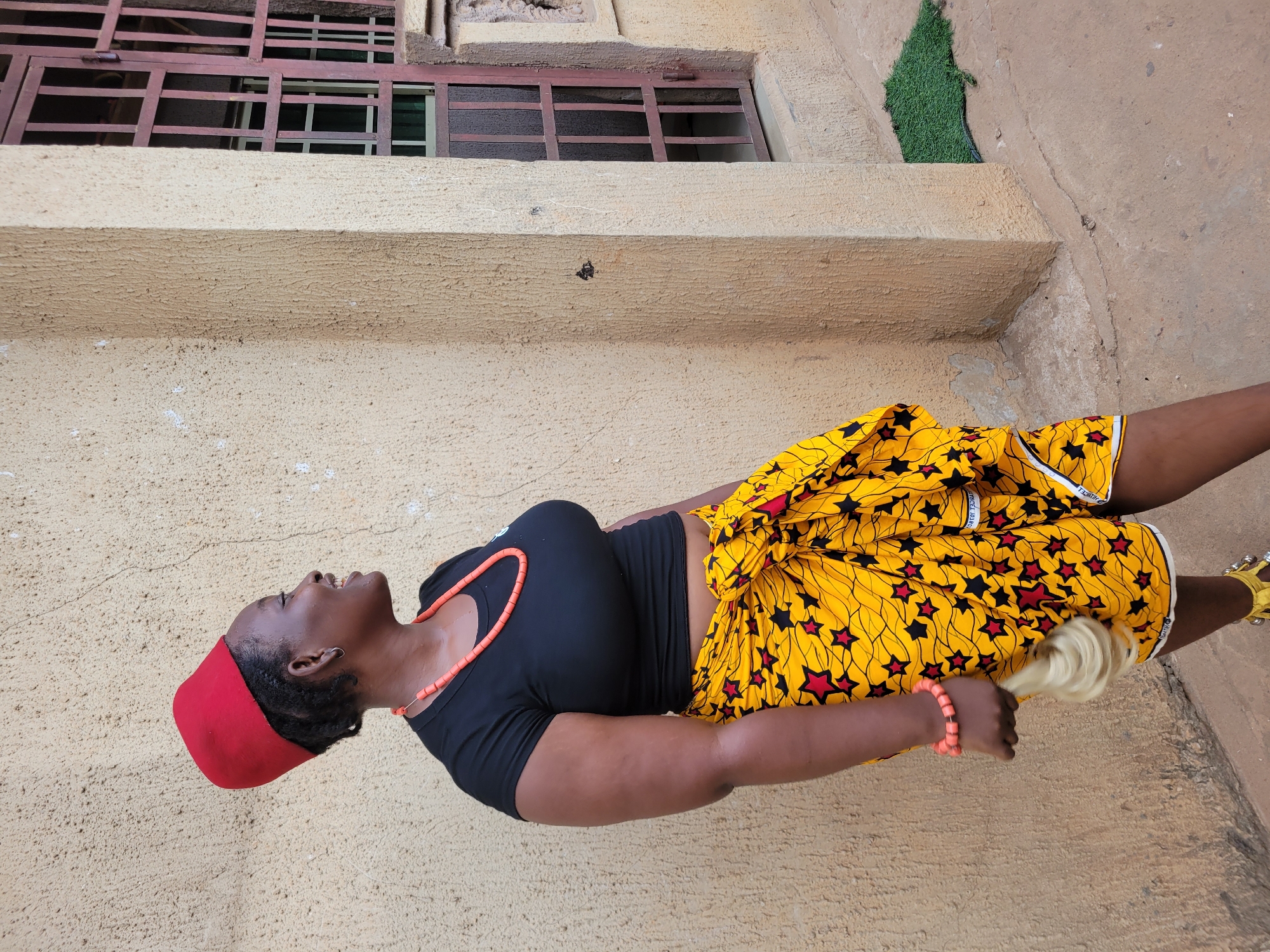 A woman wearing a red Igbo chieftaincy hat, a black top, and a yellow wrapper skirt with black and red stars is standing outside in front of a light brown building with a window with brown bars. She is wearing a coral necklace and bracelet and holding a white object in her hand. Green artificial grass is visible on a ledge behind her. The setting is likely in Onitsha, Nigeria.
