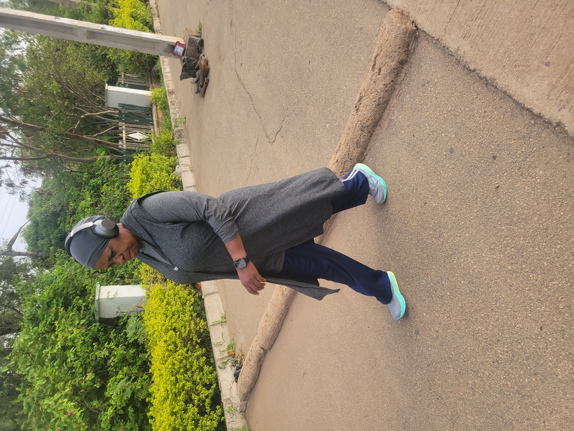 A person wearing headphones, a dark head covering, a gray long-sleeved shirt, a long gray tunic-style garment, dark blue athletic pants, and gray sneakers with light blue accents is standing on a paved road. The person is looking down. The location appears to be in Gura Top, Nigeria. To the left of the person is a small retaining wall and a fence surrounded by lush green foliage. A utility pole is visible in the background. The road appears to be cracked in some areas. Next to the utility pole is a collection of discarded items, including what appears to be pieces of leather or cloth and broken branches.