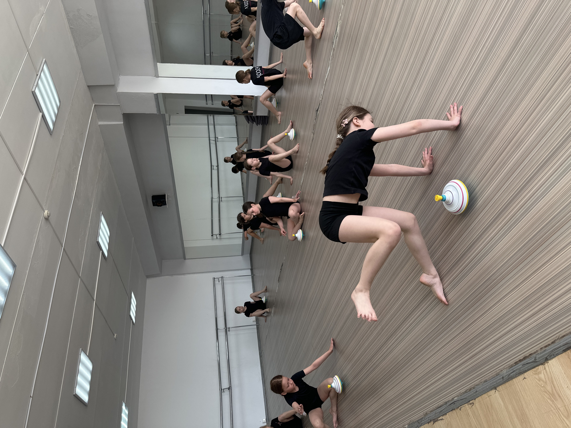 A group of approximately ten young girls, all dressed in black shirts and shorts, are performing a dance exercise in what appears to be a dance studio. The room is well-lit with rectangular ceiling lights. A large mirror runs along the length of the wall, reflecting the dancers and the room. Each girl is positioned near a spinning top toy. One girl is nearest to the camera and she stretches her arms. The location is unknown.