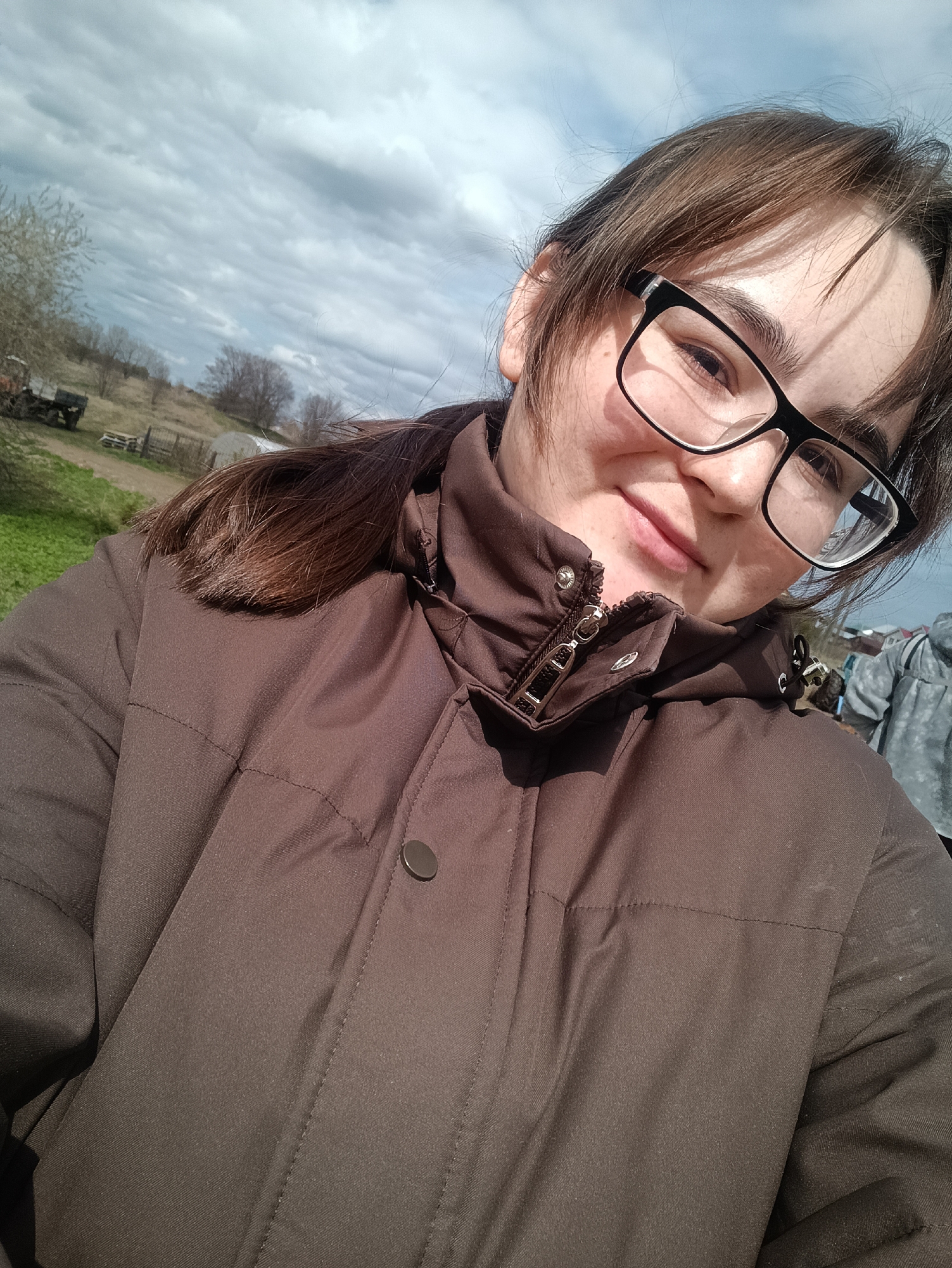 A close-up selfie captures a young person outdoors in Barda, Russia, during the daytime. The subject, who appears female, has medium-brown hair with bangs, and is wearing black-framed glasses and a dark brown, possibly padded, jacket. Her head is tilted slightly, and she looks directly at the camera with a subtle smile or smirk, conveying a relaxed and pleasant mood.

The background reveals a rural or semi-rural landscape under a partially cloudy sky, with distinct cloud formations and glimpses of blue, suggesting variable weather. The ground is covered with emerging green grass, and sparse, bare trees indicate early spring. In the distance, an old, possibly decommissioned, truck or vehicle is visible, along with a wooden fence and what appears to be a small structure like a shed or greenhouse. A dirt path or road winds through the scene. The jacket features a prominent metal zipper on the front, partially open, revealing a bit of the collar. There is no discernible text visible in the image.