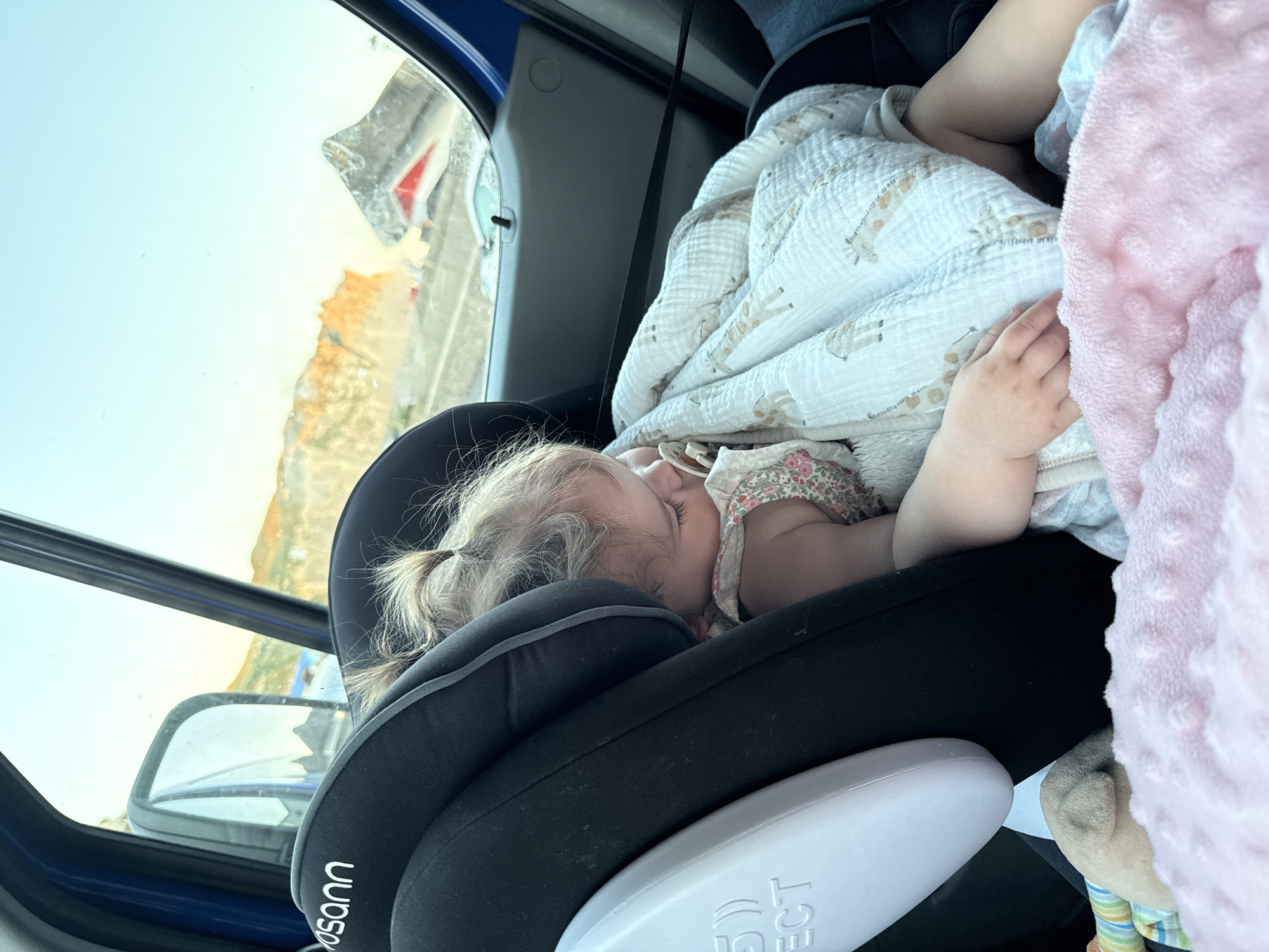 A young child is asleep in a car seat, covered by a blanket with a giraffe pattern. The child is wearing a floral print dress and has a pacifier in their mouth. The car seat is black and has the brand name "Osann" visible on it. The background shows a view through the car window of a landscape with hills and a bright sky, suggestive of a sunset. The image is cropped to focus on the child and the interior of the car. There is no indication of the location being Étretat, France in the image provided.