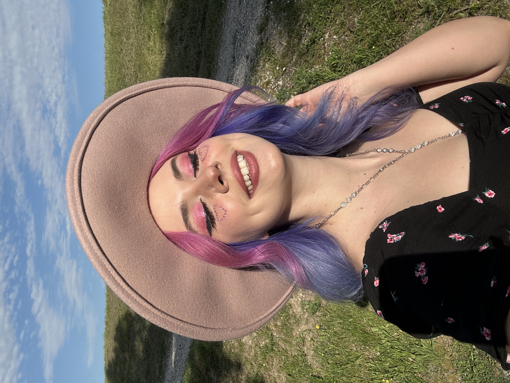 A young woman is depicted in a vibrant outdoor portrait, lying on her back on green grass, smiling broadly towards the sky. She wears a wide-brimmed, light brown felt hat. Her hair, styled with a distinct color gradient from vivid pink to purple and blue, flows around her. Her face is adorned with elaborate makeup, including bright pink eyeshadow, black eyeliner, and delicate, sparkly pink heart-shaped embellishments on her cheekbones. A silver-toned necklace featuring heart-shaped links is visible on her chest, complementing her black dress which has a subtle pink floral pattern. Her arms are bent, with one hand near her head, perhaps adjusting her hat or hair.
The setting appears to be an open, grassy area under a bright, clear blue sky with scattered white clouds, suggesting a sunny day, likely midday or afternoon. The overall mood conveyed is one of happiness, relaxation, and contentment. This serene moment takes place in an outdoor location, consistent with parks or open spaces found in the City of Zagreb, Croatia. There is no visible text in the image.