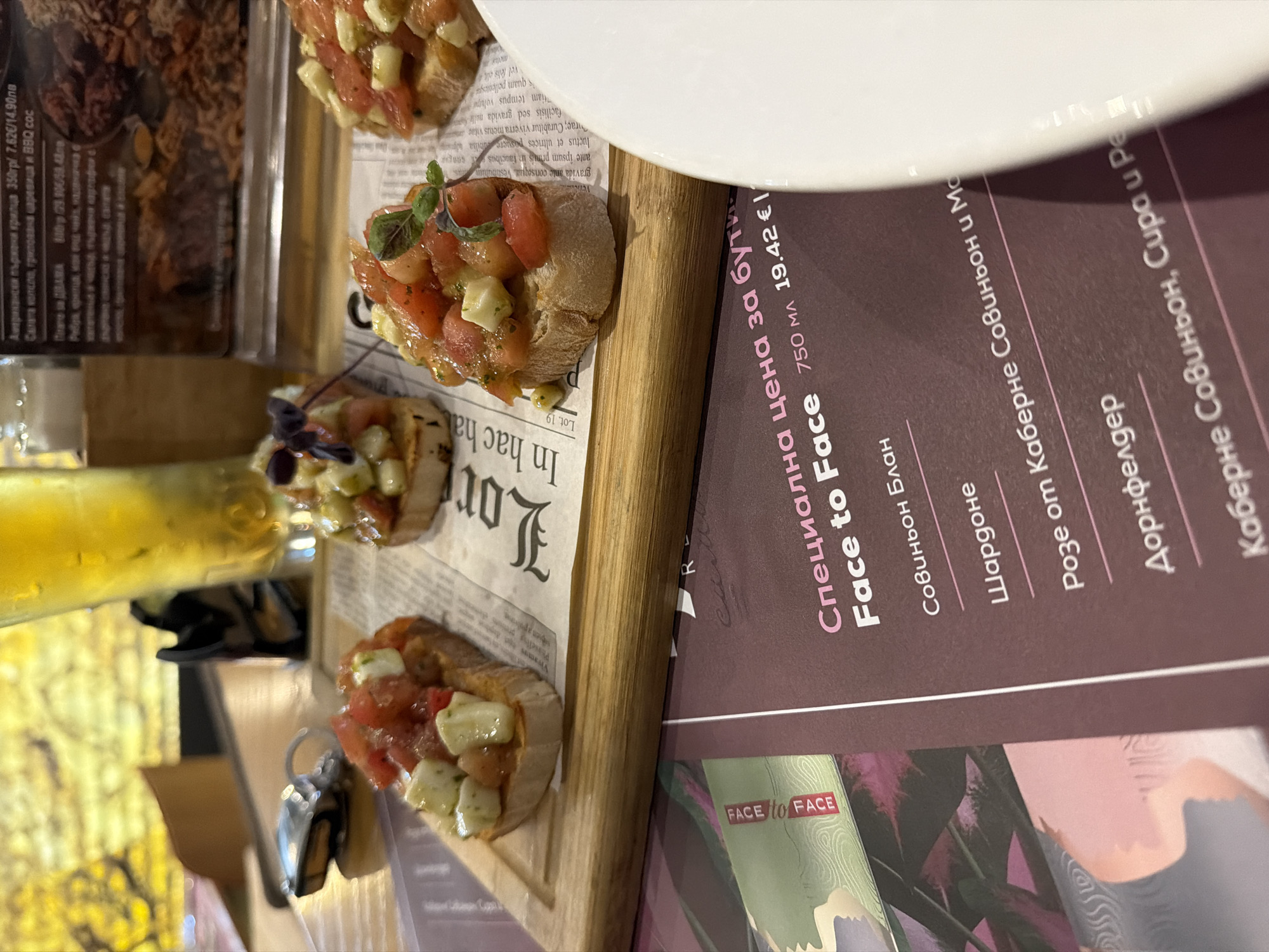 This is a close-up shot of a plate of bruschetta placed on top of a menu, likely in a restaurant. The bruschetta consists of toasted bread topped with diced tomatoes, cheese (possibly mozzarella), and herbs. A glass of yellow liquid, possibly beer or white wine, is also visible. The menu features information in Bulgarian, with the phrase "Специална цена за бути" and the brand "Face to Face." The menu lists several types of wine, including Совиньон Блан, Шардоне, Розе от Каберне Совиньон и Мо, Дорнфелдер, Каберне Совиньон, Сира и Реа. The menu also mentions other food items in Bulgarian, such as "Американски пържени крилца" (American fried wings) and "Салата колело, грилована царевица и BBQ сос" (Salad wheel, grilled corn, and BBQ sauce). The location could be inferred as Plovdiv, Bulgaria, based on the use of the Bulgarian language.