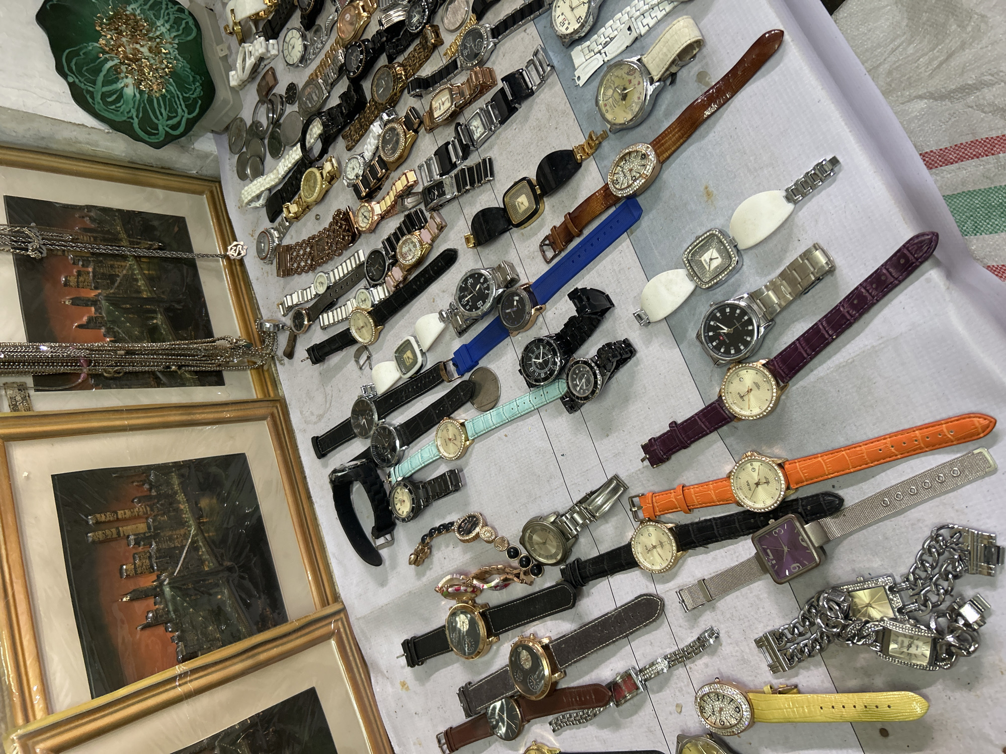 The image displays a collection of wristwatches and framed artwork, possibly at a market stall. The artwork features cityscapes, with one prominently showing the Brooklyn Bridge at night. Several watches are laid out, showcasing a variety of styles, band materials (leather, metal), and face designs. Some watches have decorative elements like rhinestones. In the background, there are necklaces and a decorative green object with gold accents. The scene appears to be indoors, with a table covered in a light-colored fabric. There are no people visible in the image. The items are arranged in a somewhat disorganized manner, suggesting a retail or flea market setting. The overall impression is one of varied goods for sale.