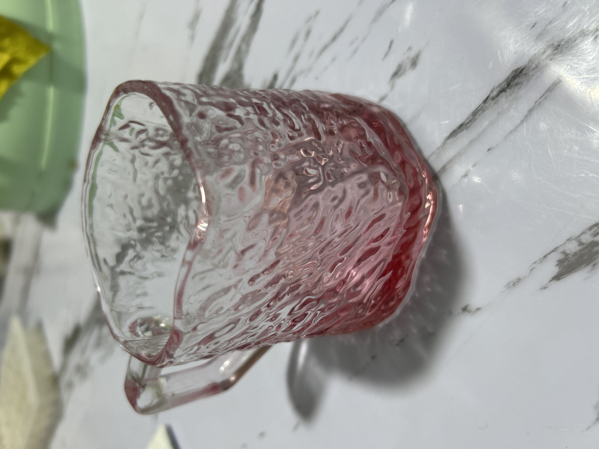 An extreme close-up shot focuses on a small, textured glass mug with a pinkish hue. The mug has a handle and is partially filled with a red liquid. It sits on a white surface with a marble-like pattern. The background is blurred and shows hints of other objects, including what appears to be a light green container. The lighting appears to be natural or from an overhead source, casting a shadow from the mug to the left. There are no people visible in the image, and the scene is static. The specific location in Дедемель, Kyrgyzstan cannot be determined from the visual information provided in the image.