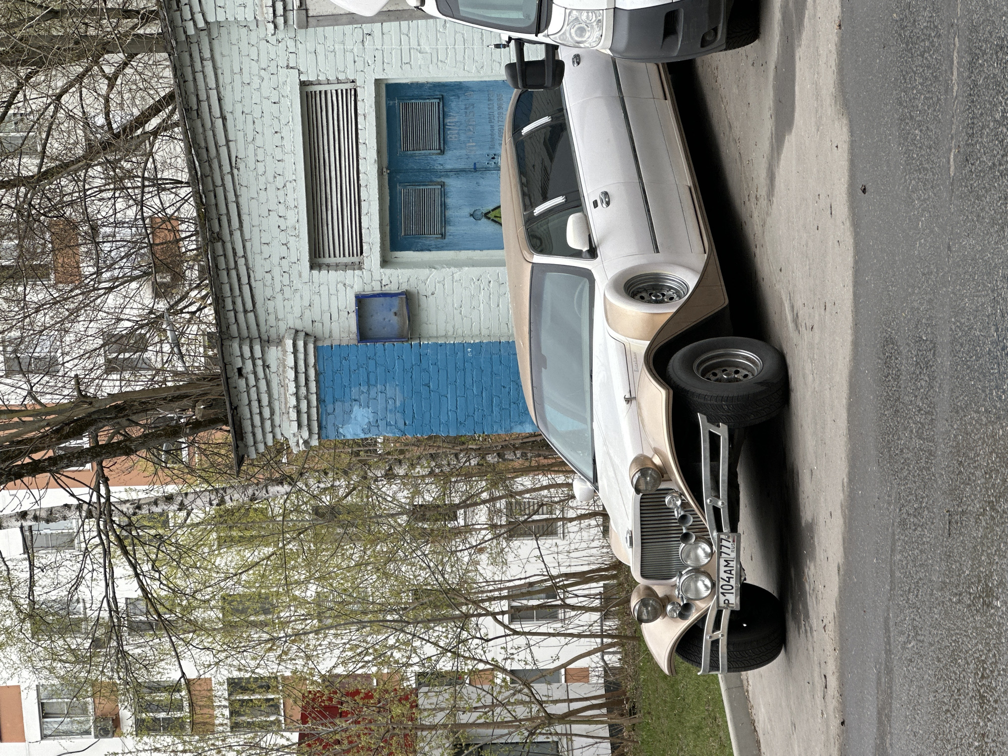 The image shows a vintage-style car parked on a street in what appears to be an urban area. The car is two-toned, white and beige, with a prominent grill and multiple headlights. It has a license plate with the designation "P 104AM 777 RUS", indicating registration in Russia. The area code "777" is associated with Moscow. The car is parked next to a brick building with blue painted doors and vents. There is a white van partially visible behind the vintage car. Trees with bare branches are in the background, and other buildings are partially visible. The street surface is asphalt. The image is taken in daylight.