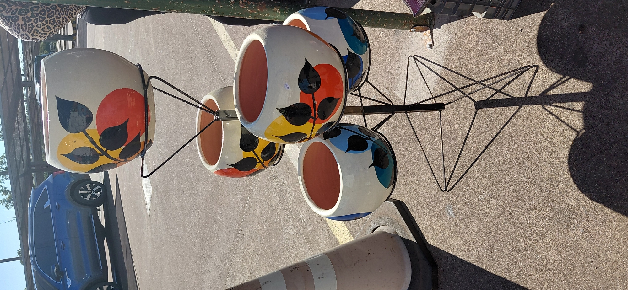 A tall, multi-tiered metal stand holds four decorative ceramic planters. The planters are light-colored with a glossy finish and feature a repeating abstract pattern of stylized leaves and circles in black, red, yellow, and blue. The top planter is the largest, with three smaller planters arranged below it in a triangular formation. The stand is black and has a geometric base.
In the background, a blue SUV is parked on asphalt. To the right, a portion of a person wearing a leopard-print top and black pants is visible. A beige traffic cone with diagonal stripes stands to the left of the planters. The scene appears to be outdoors, possibly in a market or parking lot, with a paved surface and some vegetation visible in the distance. The lighting suggests it is daytime, with strong shadows cast by the planters and the stand. The location is identified as Encarnacion, Paraguay.