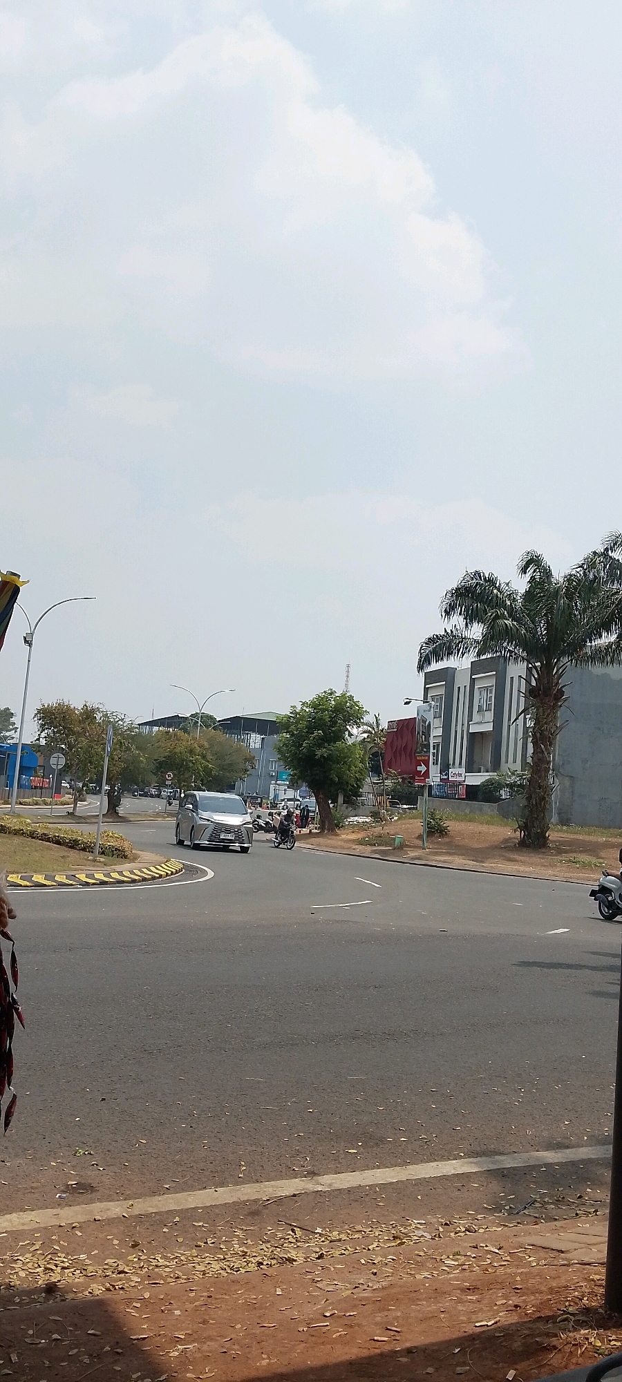 The image depicts a street scene in Gunung Putri, Indonesia, under a bright, partially cloudy sky. In the foreground, a two-lane asphalt road is visible, with a dirt shoulder to the right, littered with fallen leaves and debris. A white line marks the edge of the road.
On the road, a silver hatchback car is turning left from a side street onto the main road, which curves to the right. A motorcycle with two riders is also in motion, passing the car on the left. Further to the right, another motorcycle is seen from the rear, moving away from the viewer.
The middle ground of the image shows a roundabout or intersection. Yellow and black striped barriers demarcate the edge of the roundabout. Several trees are scattered around the area, some with green foliage, others appearing to be losing leaves. In the background, a modern, multi-story building with a gray and white facade stands prominently, adorned with dark-colored window frames. A tall palm tree with fronds is situated to the right of this building. Beyond these structures, more buildings and a tall radio tower are visible against the sky. A red sign with white text and an arrow is mounted on a pole. On the far left of the image, a portion of a yellow and blue banner hangs, attached to a lamppost. The overall environment suggests a well-trafficked urban or suburban area.