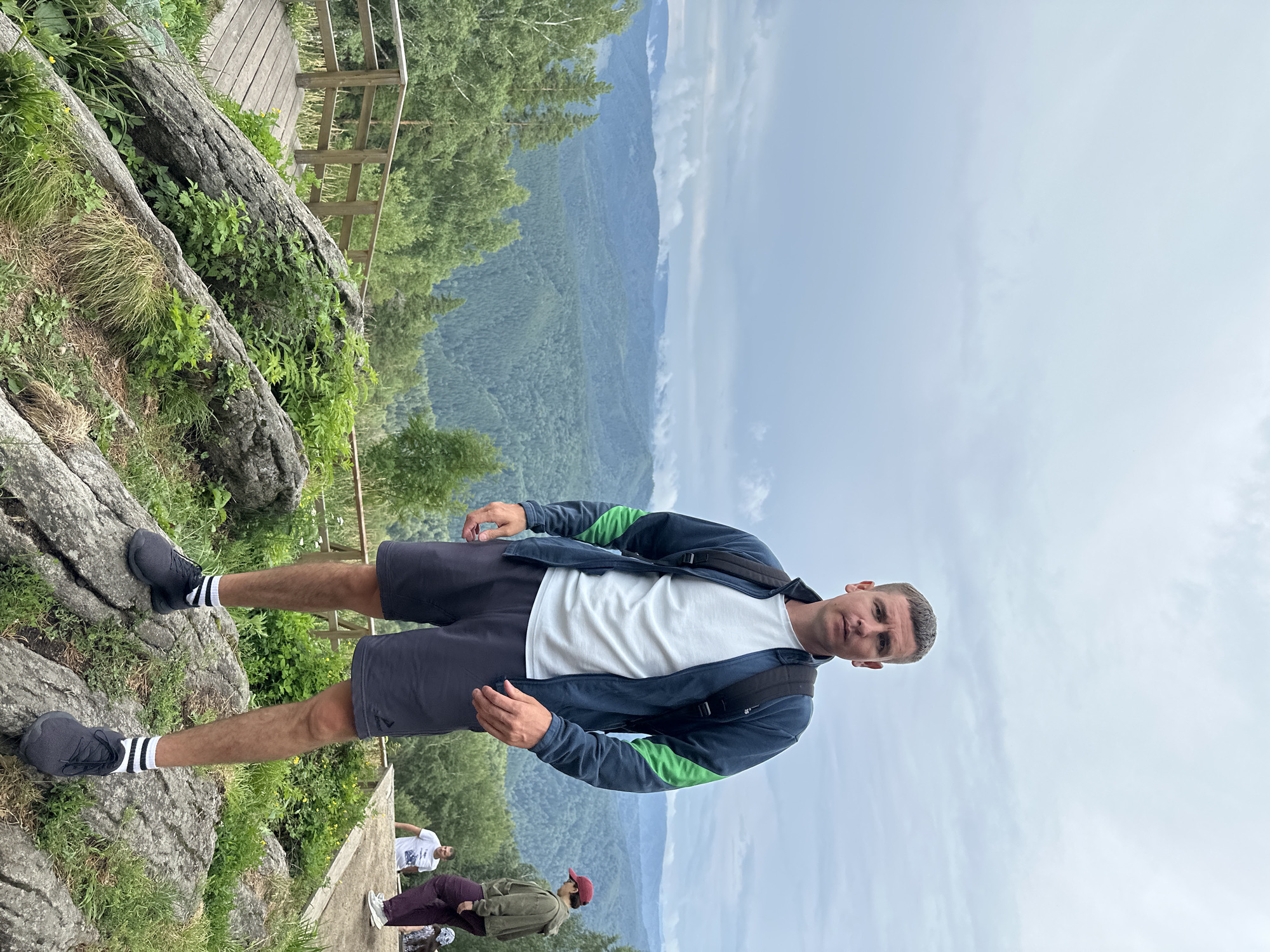 A man is standing on a rocky outcrop overlooking a vast, verdant mountain range under a cloudy sky. He is wearing a dark blue jacket with green accents, a white t-shirt, dark shorts, black athletic shoes with white-striped socks, and a black backpack. His short, brown hair is neatly styled. The man is facing the camera with a neutral expression.
In the background, to the right, two other individuals are visible. One is wearing a green hooded jacket and dark pants, with a red baseball cap. The other is wearing a white t-shirt and light-colored pants. They appear to be walking on a wooden path or viewing platform.
The foreground consists of large, weathered rocks covered in green vegetation, including grasses and small leafy plants. A wooden railing and a wooden walkway are visible to the left, suggesting a designated viewing area or trail. The mountains in the distance are covered in dense evergreen forests, with layers of mist or clouds obscuring the furthest peaks.
The overall scene depicts a natural mountain landscape, likely a scenic viewpoint, possibly in or near the city of Belokurikha, Russia, known for its mountainous terrain and natural beauty. The weather appears overcast, contributing to the muted colors of the landscape.