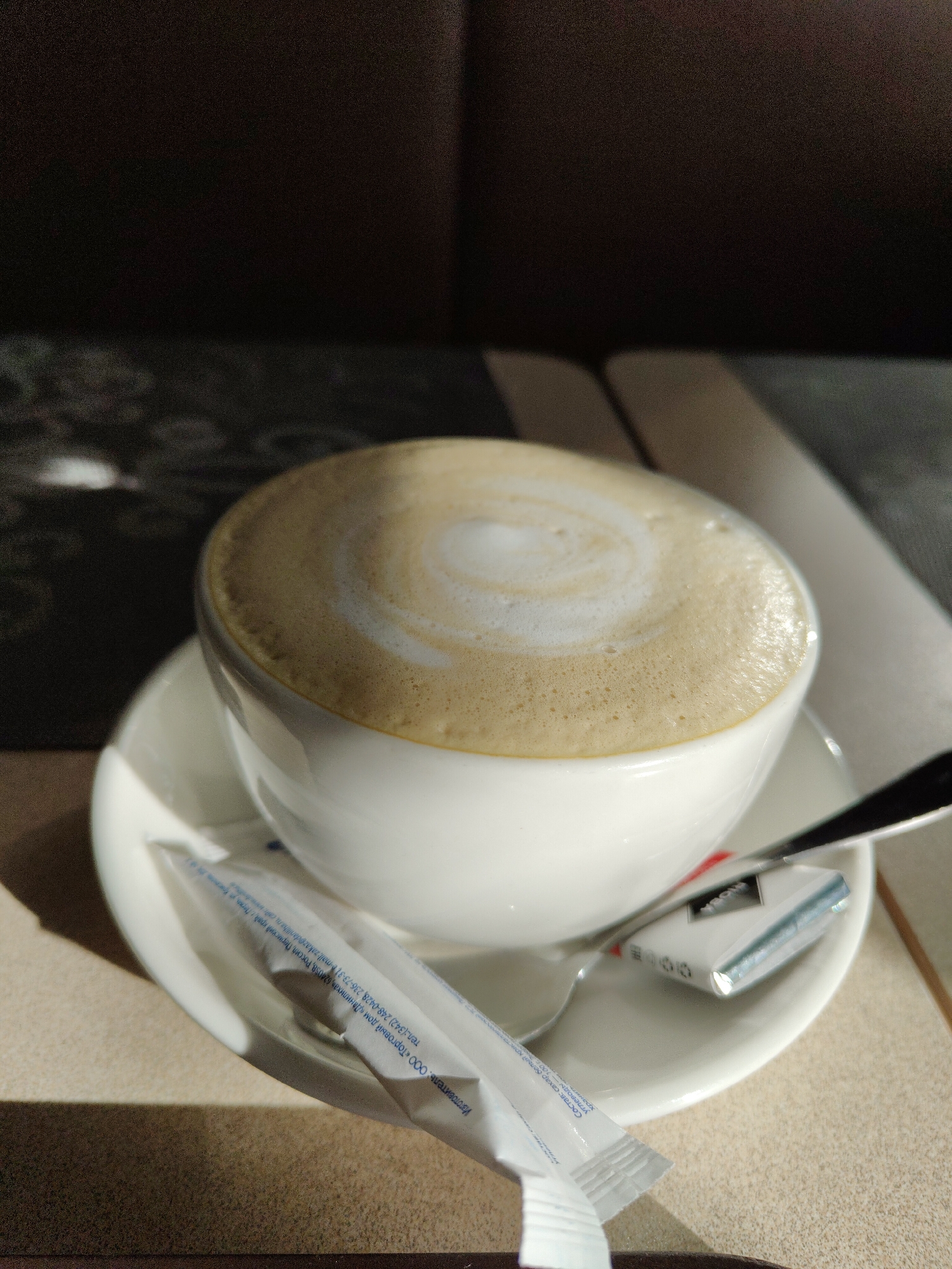 A close-up shot depicts a cup of cappuccino on a saucer, accompanied by a sugar packet and a spoon. The cup is white, overflowing with foamy cappuccino that features a circular swirl design on top. The saucer is also white and holds a white sugar packet and a small spoon. The background includes a dark, patterned surface behind the cappuccino and a light beige surface beneath it. The scene is lit with a warm, strong light. The sugar packet shows Russian text: “Изготовитель: ООО «Торговый дом «Данилиха”, 624058, Россия, Перческий край Перва тел.: (342) 248-0428, 236-73-31-mail: zakaz@danilihan cale Состав: сахар белый кристалличней углеводы хранить 100 г".