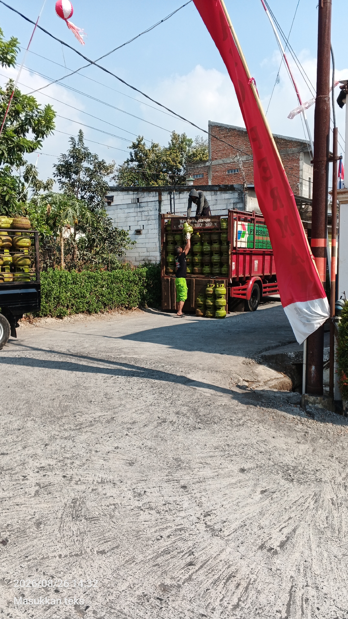 In Jeru, Indonesia, two individuals are engaged in the unloading of gas cylinders from a truck. The scene depicts a sunny day with a clear sky. A red truck is parked on a paved road, loaded with numerous green gas cylinders. One person, wearing a dark hoodie, is positioned on top of the truck, actively handling the cylinders. Another person, dressed in a black t-shirt and bright green shorts, stands on the ground next to the truck, receiving a cylinder. To the left of the truck, a black pickup truck is also visible, with a load of gas cylinders stacked on its flatbed. In the background, a white brick building and some trees are present, along with a tall pole adorned with a red and white flag. The ground in the foreground is a gray, textured concrete surface. The image also contains a timestamp and text overlay that reads "2023/08/26 14:32 Masukkan teks".