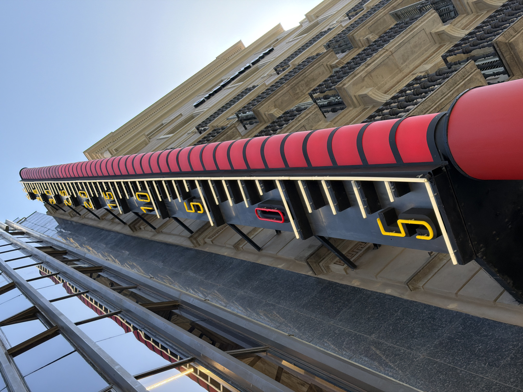 The image shows a tall, red and black thermometer on a street in Barcelona, Spain. The thermometer is positioned next to a modern building with a glass facade and an older building with balconies. The thermometer has yellow numbers and markings, and a red indicator that appears to be at a high temperature. The sky is clear and blue. The perspective is low-angle, looking up at the thermometer and the buildings. There are no people visible in the image.
