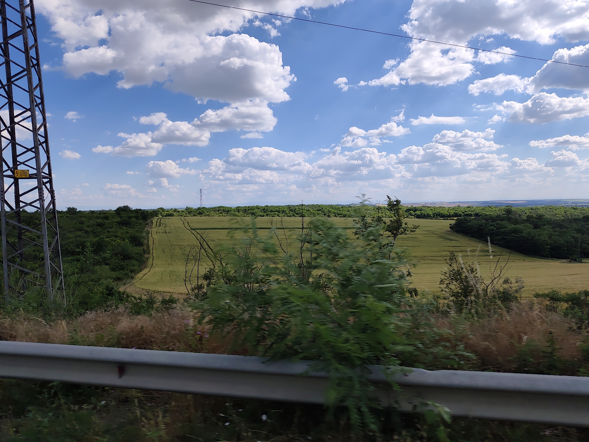 The image depicts a wide, rural landscape under a bright, cloudy sky. In the foreground, a metal guardrail runs horizontally across the bottom of the frame, partially obscured by dry grasses and green foliage. To the left, a large, dark metal lattice tower stands prominently, its structure detailed with crisscrossing beams. A yellow warning sign is affixed to the tower.
Beyond the immediate foreground, the scene opens up to a vast, green field with visible tire tracks suggesting agricultural activity. This field is bordered by dense, dark green trees that form a natural boundary. In the distance, the landscape rolls gently, with more trees and fields stretching towards the horizon. The sky is a vibrant blue, dotted with fluffy white cumulus clouds, indicating fair weather. The lighting suggests it is daytime, likely late morning or early afternoon.
The overall impression is one of peaceful, open countryside. There are no people visible in the image. The setting appears to be a rural area in Zgalevo, Bulgaria, as indicated by the context. The image captures a moment of natural beauty and agricultural expanse, likely taken from a moving vehicle due to the slight blur in the foreground vegetation.