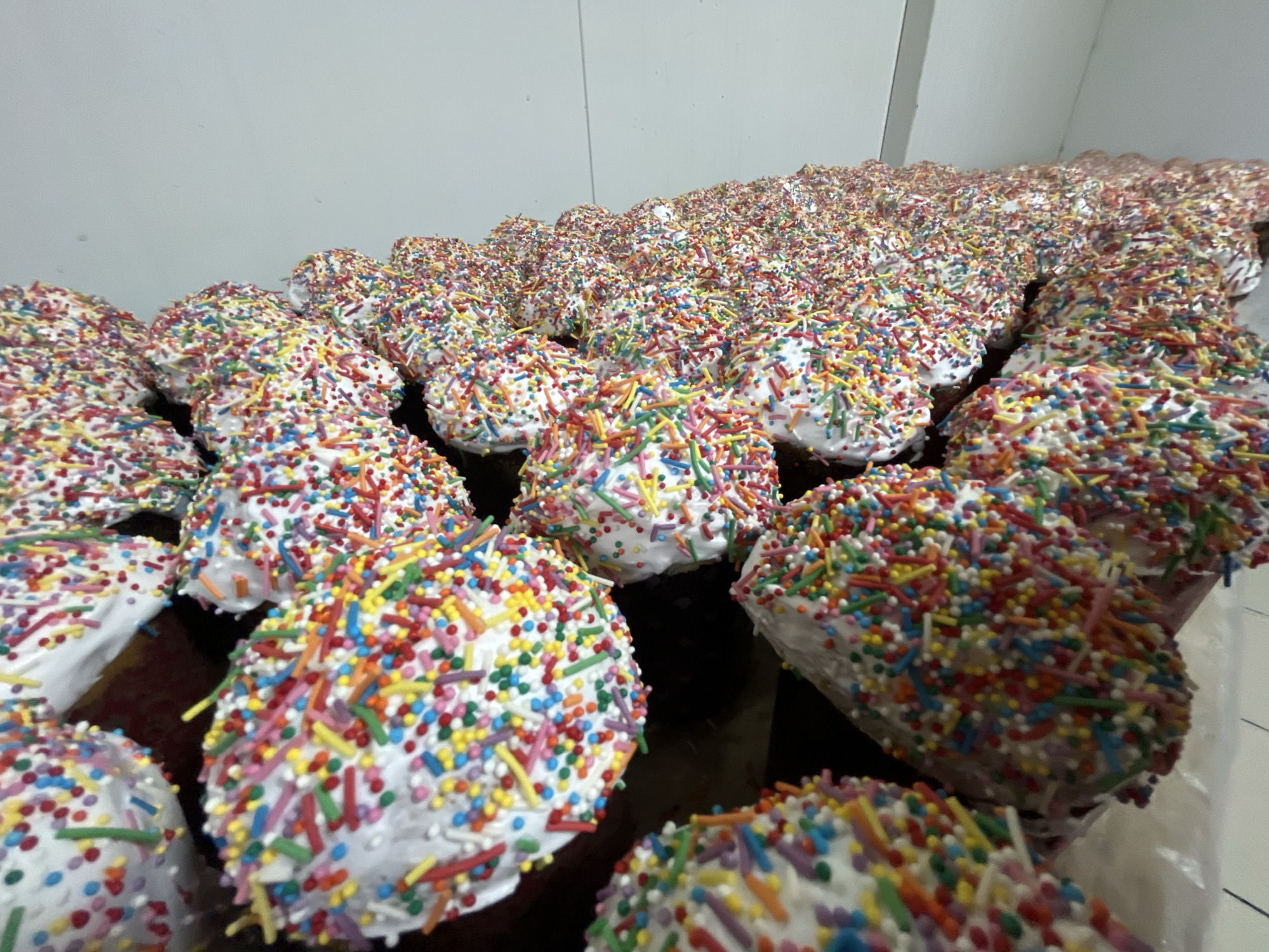 The image depicts a large quantity of baked goods, likely Kulich cakes, arranged on a surface. The cakes are covered in white icing and topped with multicolored sprinkles. The background suggests a commercial kitchen or food preparation area, possibly in a bakery. The lighting is bright and artificial. There are no people visible in the frame. The image lacks enough detail to confirm its location as Novopokrovskaya, Russia.