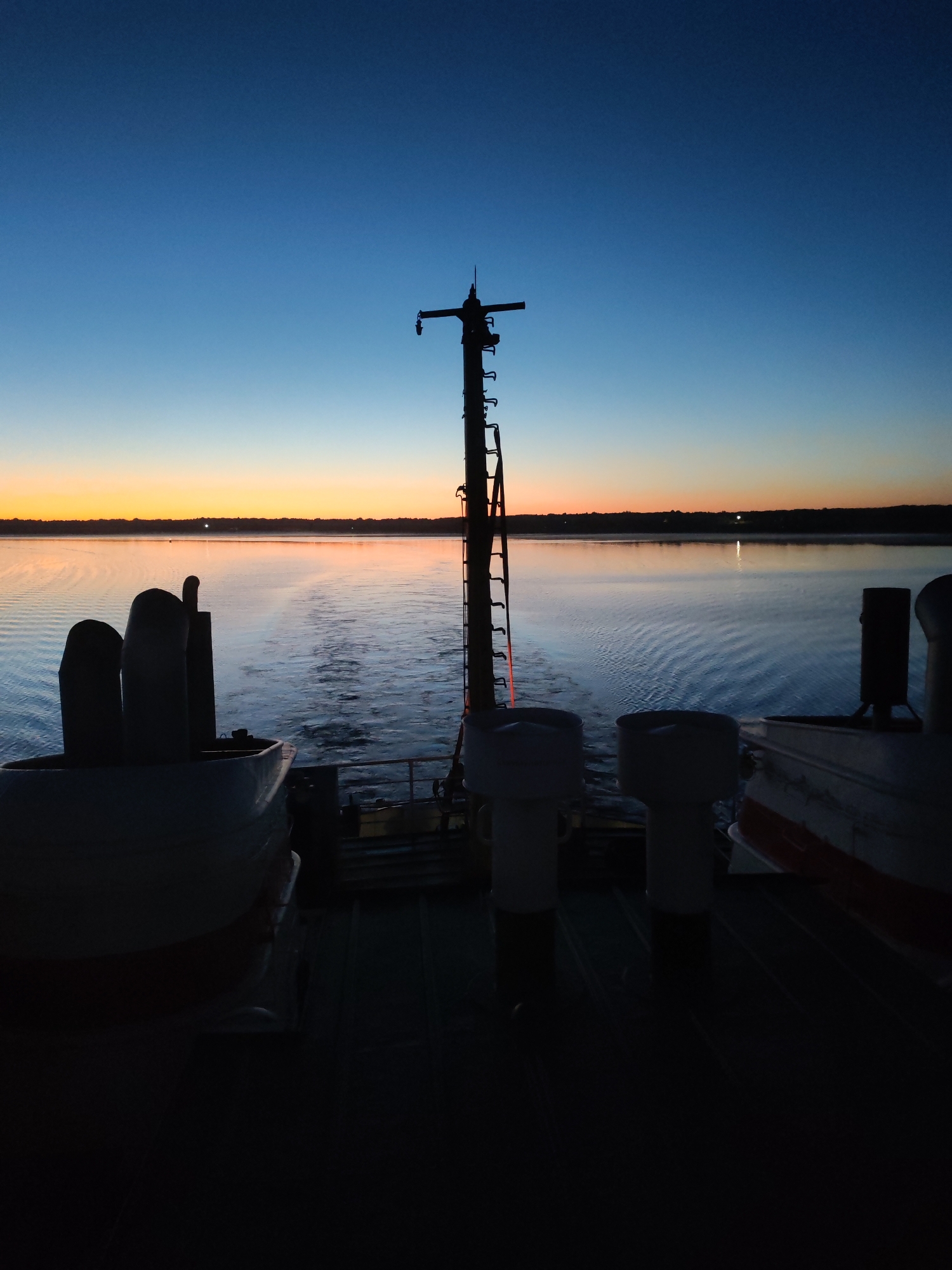The image captures a twilight or dawn scene viewed from the stern of a vessel navigating a body of water near the city of Раменье, Russia. The sky exhibits a gradient from deep blue overhead to orange and yellow at the horizon, indicative of sunrise or sunset. This atmospheric light is partially reflected on the calm water surface. A dark, indistinct shoreline or landmass is visible on the distant horizon, featuring a few faint artificial lights. In the immediate foreground, the stern of the vessel is rendered in deep silhouette. A prominent, tall, dark mast-like structure equipped with a ladder and various components rises vertically near the center. To the left, two dark, curved, tubular structures, likely ventilation or exhaust elements, are visible on the deck. On the right, additional silhouetted boat structures and railings are present. A distinct wake trails behind the vessel, indicating its movement through the water. No individuals are visible in the frame.