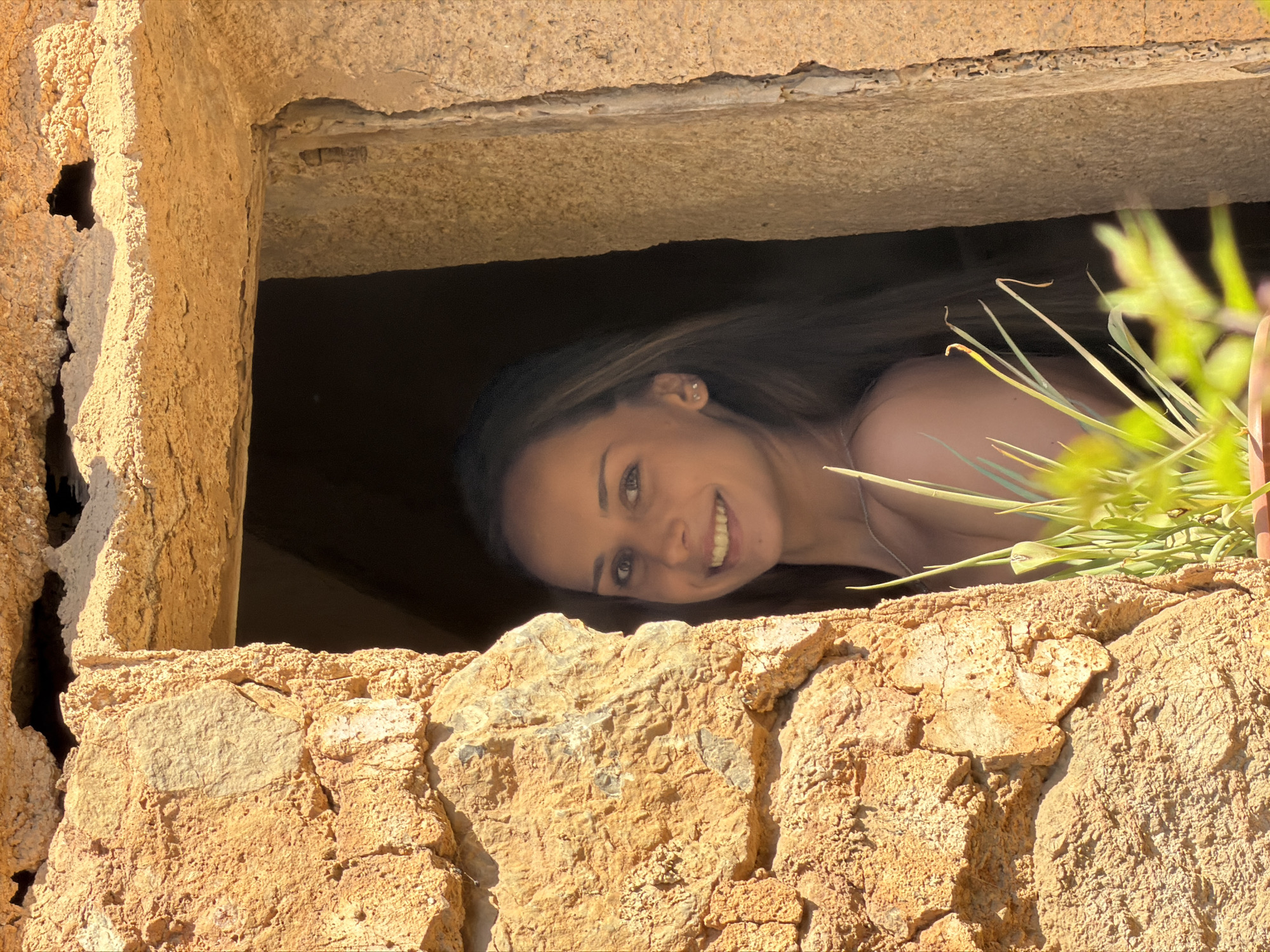 A young woman is looking out from a window in a stone structure. The structure appears old and weathered. The location is likely Bunyola, Spain, given the request for that context. The woman has long hair and is smiling. Some green foliage is visible to the right of the window. The inside of the structure is dark.
