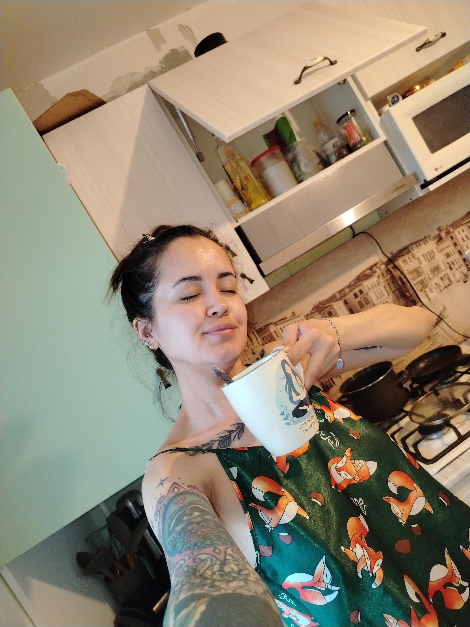 The image captures a selfie of a young woman in a kitchen, exuding a sense of calm and enjoyment. Her eyes are closed, and a subtle smile plays on her lips as she holds a white mug with a spoon inside to her mouth. Her dark hair is styled back, and she sports multiple ear piercings and a nose ring. Her left arm and shoulder are extensively covered in detailed tattoos. She is dressed in a dark green sleeveless top patterned with whimsical orange foxes and small brown acorns.
The white mug features a blue and white graphic of a meditating woman, with Cyrillic text partly visible that reads "ДЛЯ ЛЮБВИ НЕ НУЖЕН" (DLYA LYUBVI NE NUZHEN), translating to "FOR LOVE YOU DON'T NEED."
The background shows a home kitchen setting. Light-colored upper cabinets line the wall; one is open, revealing various jars and bottles. Adjacent to it, a white LG microwave is visible. Below the cabinets, a metallic range hood, partially showing the brand "DAGINA," is positioned above a gas stove with several pots and pans. A decorative backsplash behind the stove features a detailed illustration of a European cityscape, possibly Venice, with historical buildings and waterways. Notably, the wall above the kitchen cabinets appears unfinished or in disrepair, with visible patches of exposed plaster or peeling paint. A light green wall borders the left side of the frame. The scene suggests a tranquil moment, likely in the morning or early day, within a residence in Noviy, Russia.