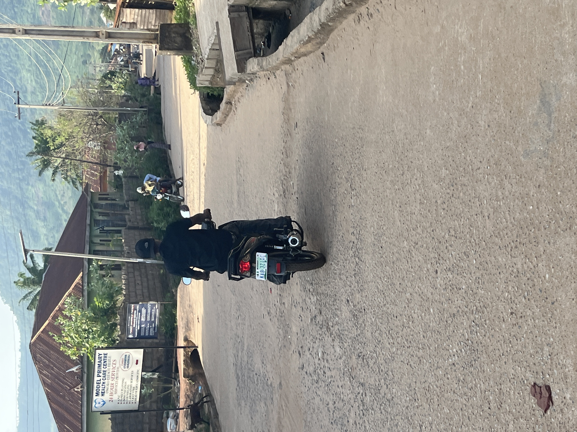 A person is riding a motorcycle down a dusty, unpaved road in a village. The motorcycle is dark colored with a license plate that reads "KAB-32LOC". The rider is wearing a black shirt and a black baseball cap. The road is lined with buildings and vegetation. In the background, there are hills with some greenery. There are other people and motorcycles visible further down the road. A sign on a building to the left advertises "MODEL PRIMARY HEALTH CARE CENTRE". A goat is visible on the left side of the road. The scene appears to be during daylight hours.