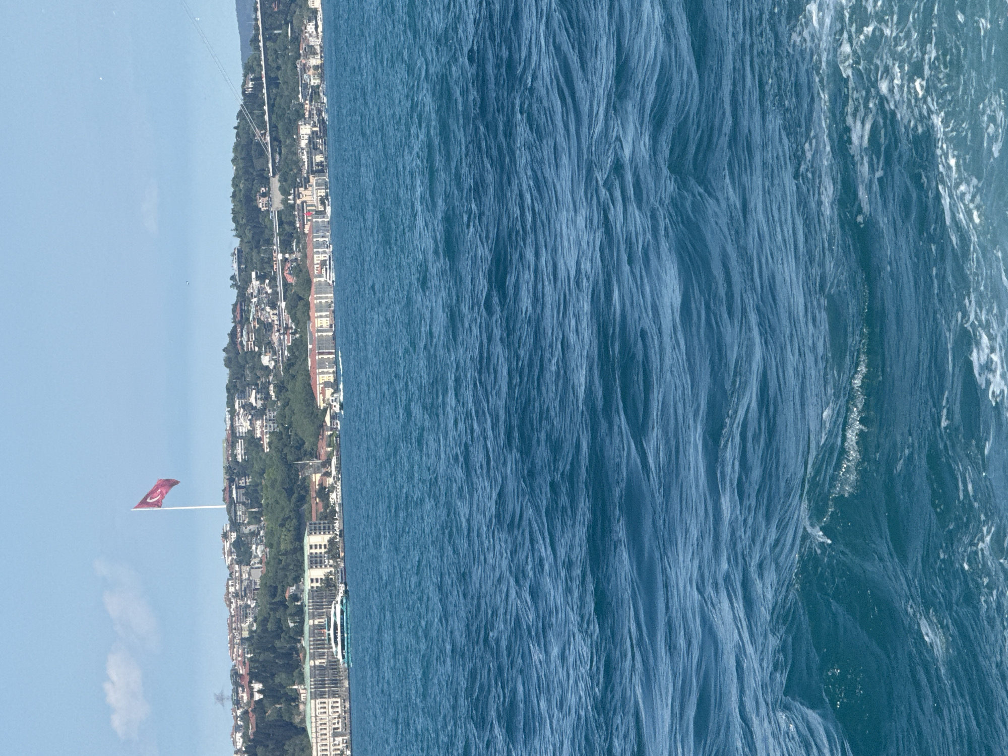 The image presents a long shot of a cityscape bordering the sea. The scene is oriented vertically, with the city appearing along the left edge and the sea occupying the right. The sky is visible in the upper-left, showing a clear, light blue sky. A Turkish flag is prominent in the center-left of the image, close to the cityscape. The water is deep blue with prominent ripples and streaks, indicating movement. Buildings of varying heights and architectural styles line the coast, interspersed with patches of green vegetation.