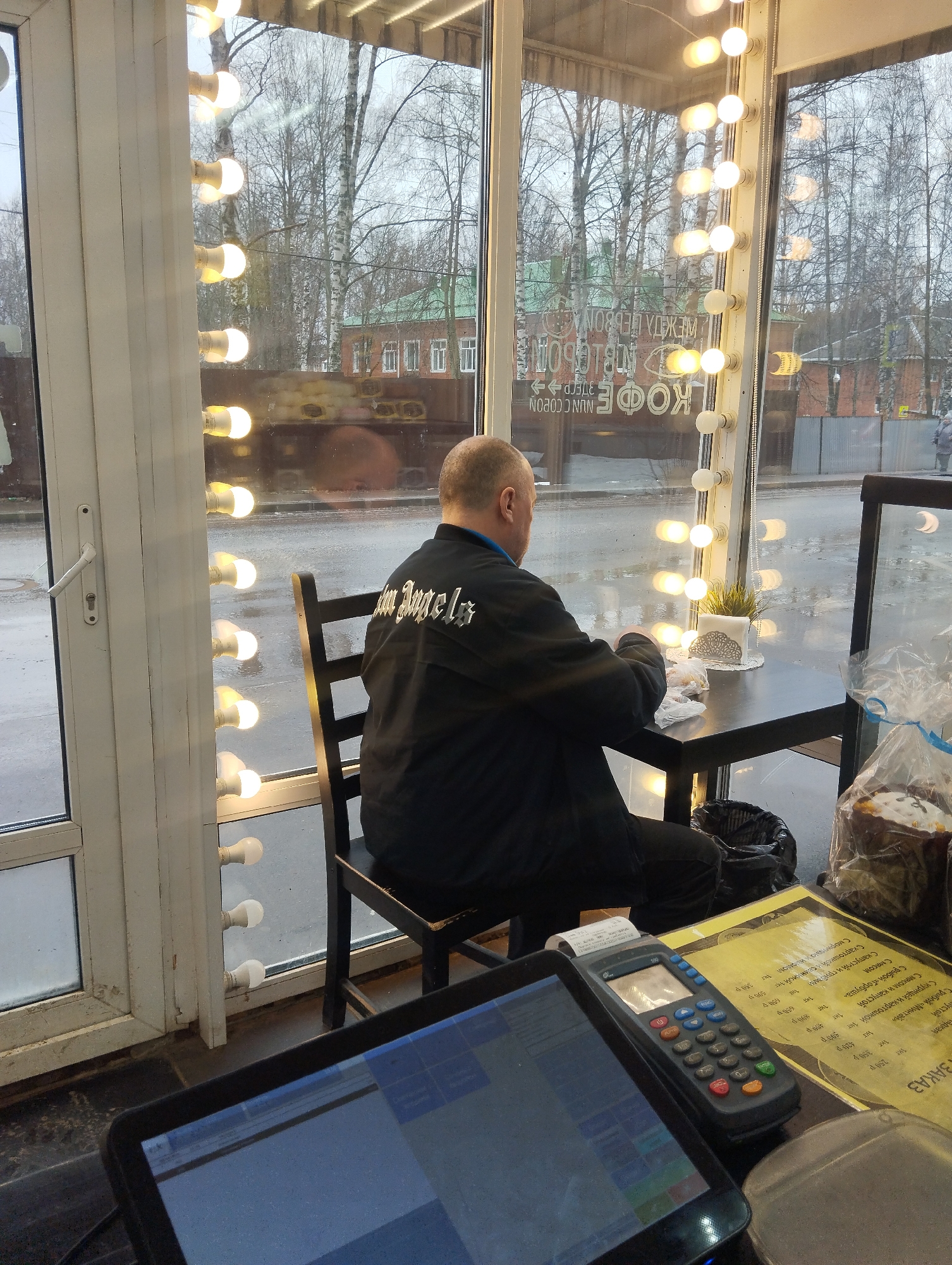 Here is a forensic description of the image:
**Scene:** A view from inside what appears to be a cafe in Votkinsk, Russia. The view is looking out a large window towards a street scene on a cloudy day.
**Subject:** A man is seated at a small black table near the window. He has a bald head and is wearing a black jacket with "Palm Angels" printed on the back. He is focused on something on the table in front of him, potentially eating.
**Objects:**
* Several vertical rows of light bulbs are positioned along the window frame, creating a lighted border.
* Outside the window, there are trees without leaves, buildings, and a glimpse of the street.
* On the table where the man is sitting, there is a small potted plant, possibly a succulent, and some items wrapped in plastic.
* A black trash receptacle is next to the man.
* In the foreground, a point-of-sale (POS) system is visible, including a payment terminal and a printed menu with prices, some items are 'мясом' 'С мясом и капустой', written in Russian.
**Environment:** The overall environment appears to be a mix of indoor and outdoor with subdued lighting due to the overcast day. The cafe seems to have a modern, clean style with a focus on the view outside.
**Action:** The man is occupying himself, possibly having a meal, inside the cafe. The presence of the POS terminal suggests the cafe is open for business.
**Location:** The presence of Russian text and architecture details suggests the location is in Votkinsk, Russia.