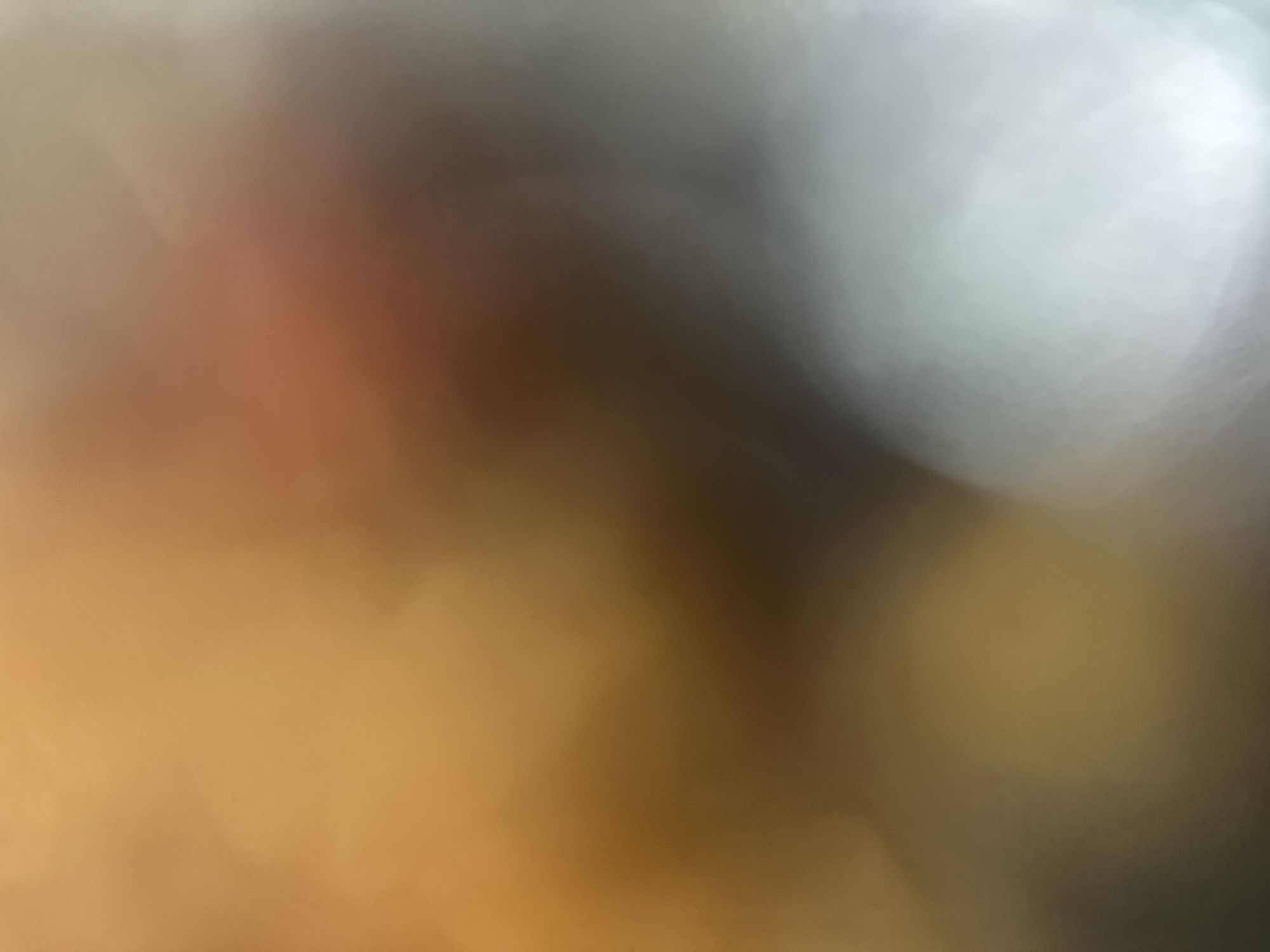 This is an abstract, out-of-focus image with a bokeh effect. The dominant colors are warm earthy tones, ranging from deep orange and brown in the lower left to lighter tan and gray tones towards the upper right. There are areas of bright, diffused light that create soft, circular blurs. The overall impression is one of diffused light and color, without any discernible subjects, objects, or a specific scene. No text is visible. The image lacks any clear indicators of time of day, weather, location, or emotions due to its abstract nature.