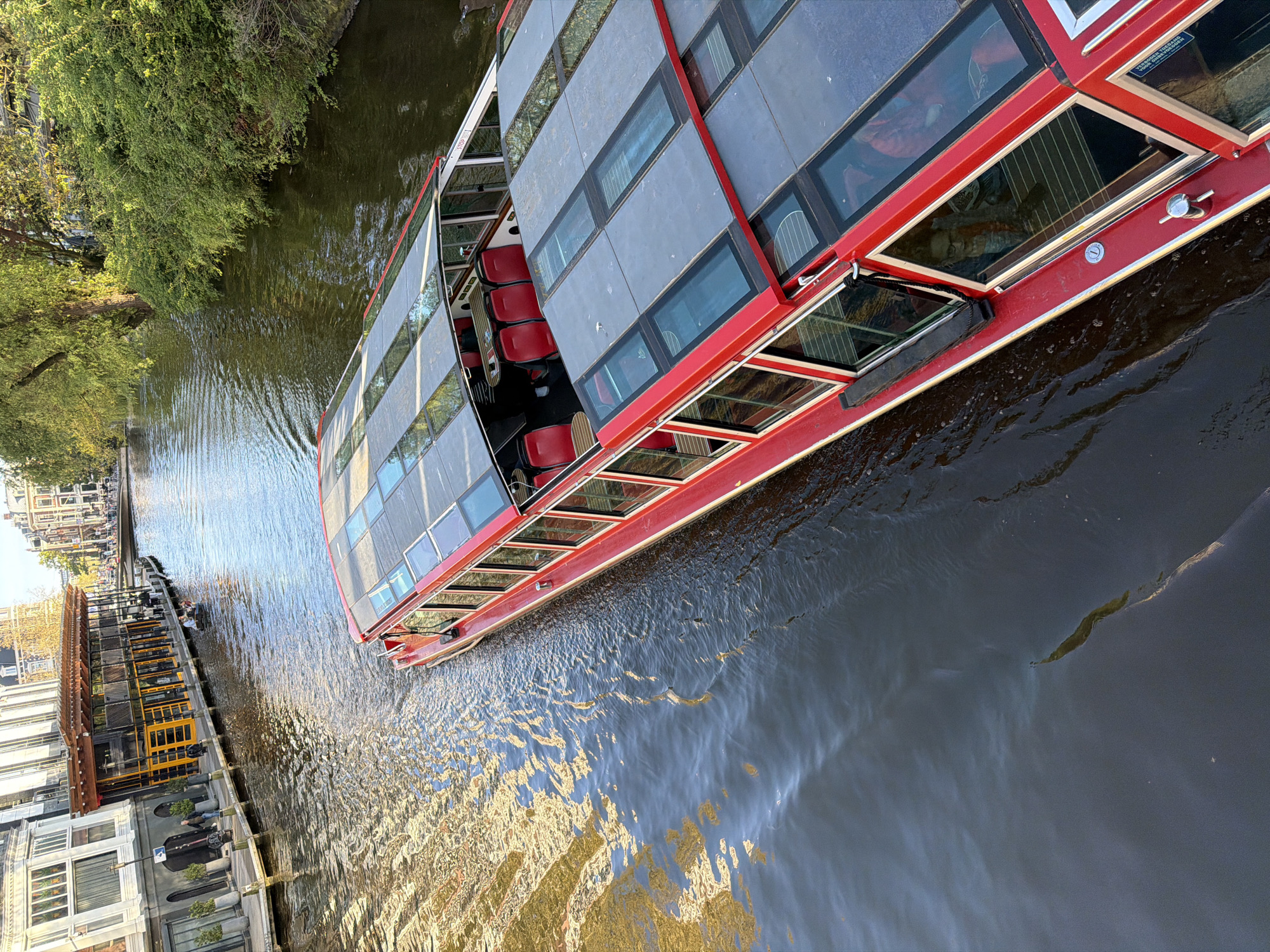 A red canal tour boat navigates a canal in Amsterdam, Netherlands. The boat, with a transparent roof and multiple windows, is partially open on one side. Rows of red seats are visible inside. The canal water is dark, with reflections of buildings and trees. On the left bank, there's a modern building with yellow accents. A tree overhangs the water. The lighting suggests it's daytime.