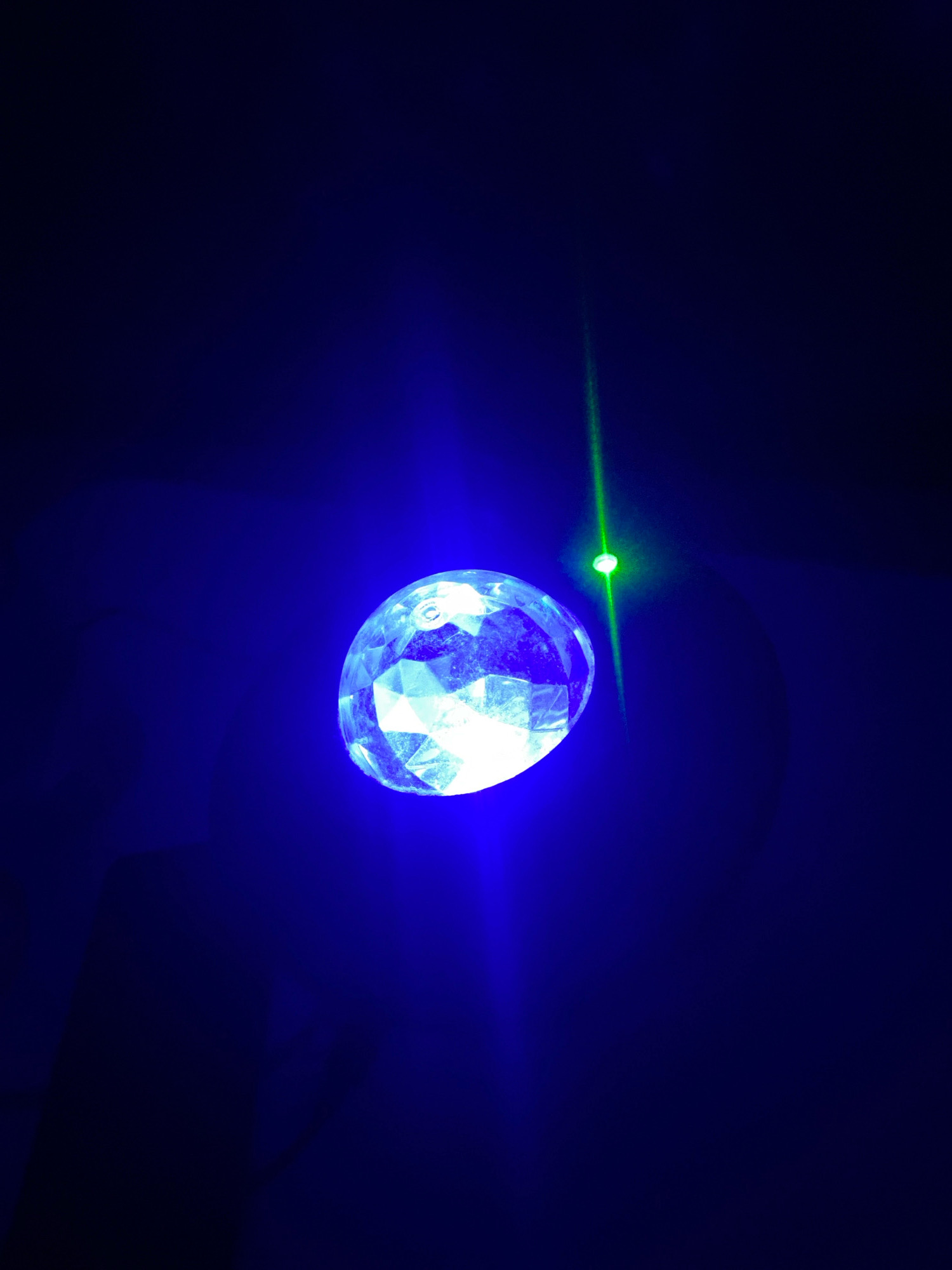 A faceted, translucent orb glows with bright white and blue light against a dark background. A thin, bright green laser beam extends from the upper right of the orb diagonally upward and slightly to the left. A small, bright green point of light is visible near the top of the laser beam. The scene is dark, with the primary light sources being the orb and the laser. The location in Klurak, Indonesia, is not discernible from the image.