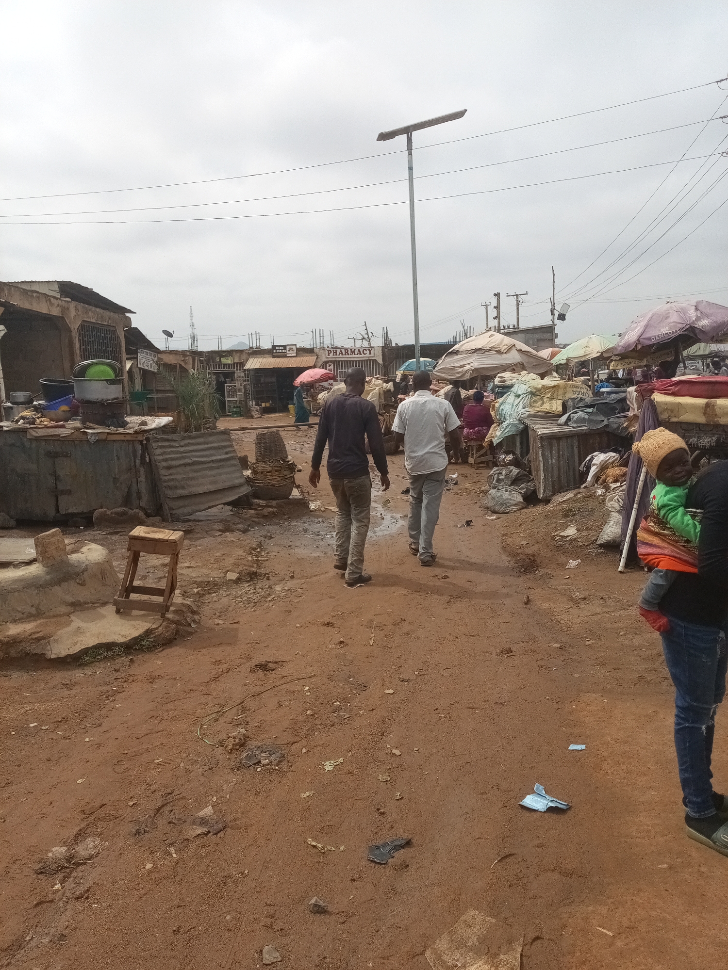 The image depicts a busy marketplace scene in Rafin Par, Nigeria. Two men are walking away from the camera on a dirt path. The man on the left is wearing a dark long-sleeved shirt and light-colored pants. The man on the right is wearing a white short-sleeved shirt and light-colored pants. Behind them, a woman is carrying a baby wrapped in a sling, with the baby wearing a yellow hat and green garment. The marketplace is filled with various stalls and vendors. Some stalls have corrugated metal roofs and sides, displaying goods such as cooking pots, baskets, and sacks of materials. Colorful umbrellas are set up to provide shade. In the background, buildings line the street, with one prominently displaying a sign for "PHARMACY." A solar-powered street lamp is visible in the distance, along with power lines crisscrossing the sky. The ground is uneven and covered with red-brown dirt, with some debris and puddles of water scattered around. The sky is overcast, suggesting a cloudy day.