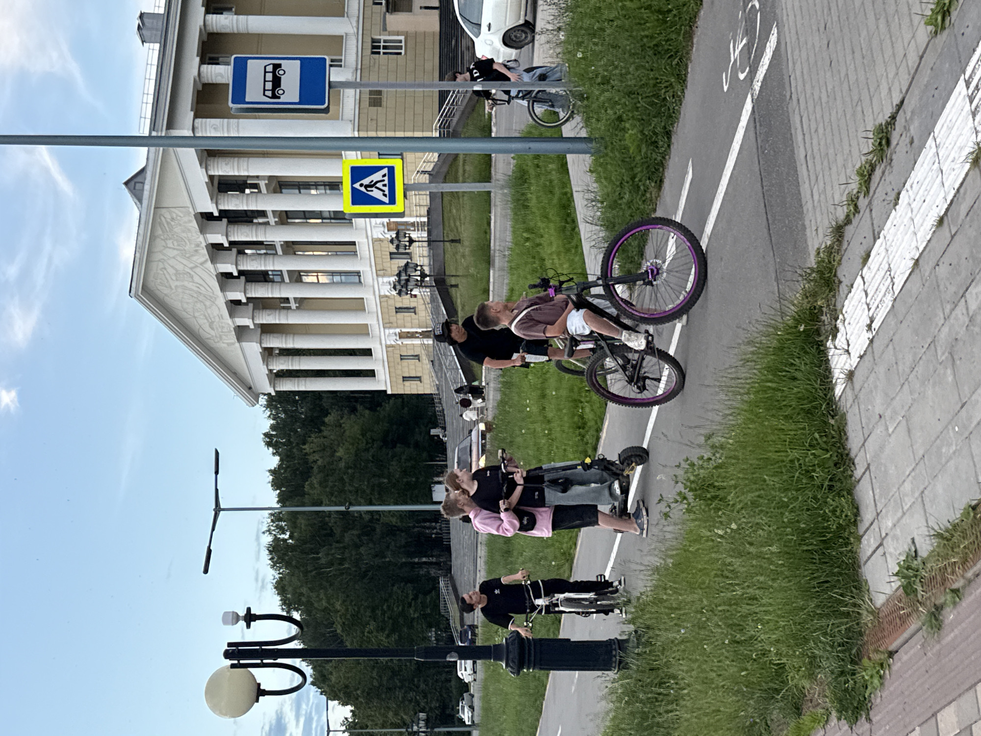 The image depicts a group of young people on a sunny day in Votkinsk, Russia. The scene is set in an urban park or promenade area with a paved path marked for bicycles, flanked by green grass and a sidewalk. In the background, a grand, classical-style building with columns and a pediment is visible. Two road signs are present: one indicating a bus stop with a silhouette of a bus, and another a yellow triangle with a pedestrian symbol inside.
Several individuals are engaged in activities related to cycling and scooters. One person is leaning against a bicycle with purple rims. Another group of three appears to be on a scooter, with two individuals standing and one possibly sitting, all looking upwards or at each other. Further down the path, another individual is seen riding a bicycle. The overall mood seems casual and relaxed, suggesting a leisurely outing. The time of day is likely afternoon, given the bright sunlight and clear sky. The presence of a building with distinct architectural features and streetlights suggests a public, well-maintained area.