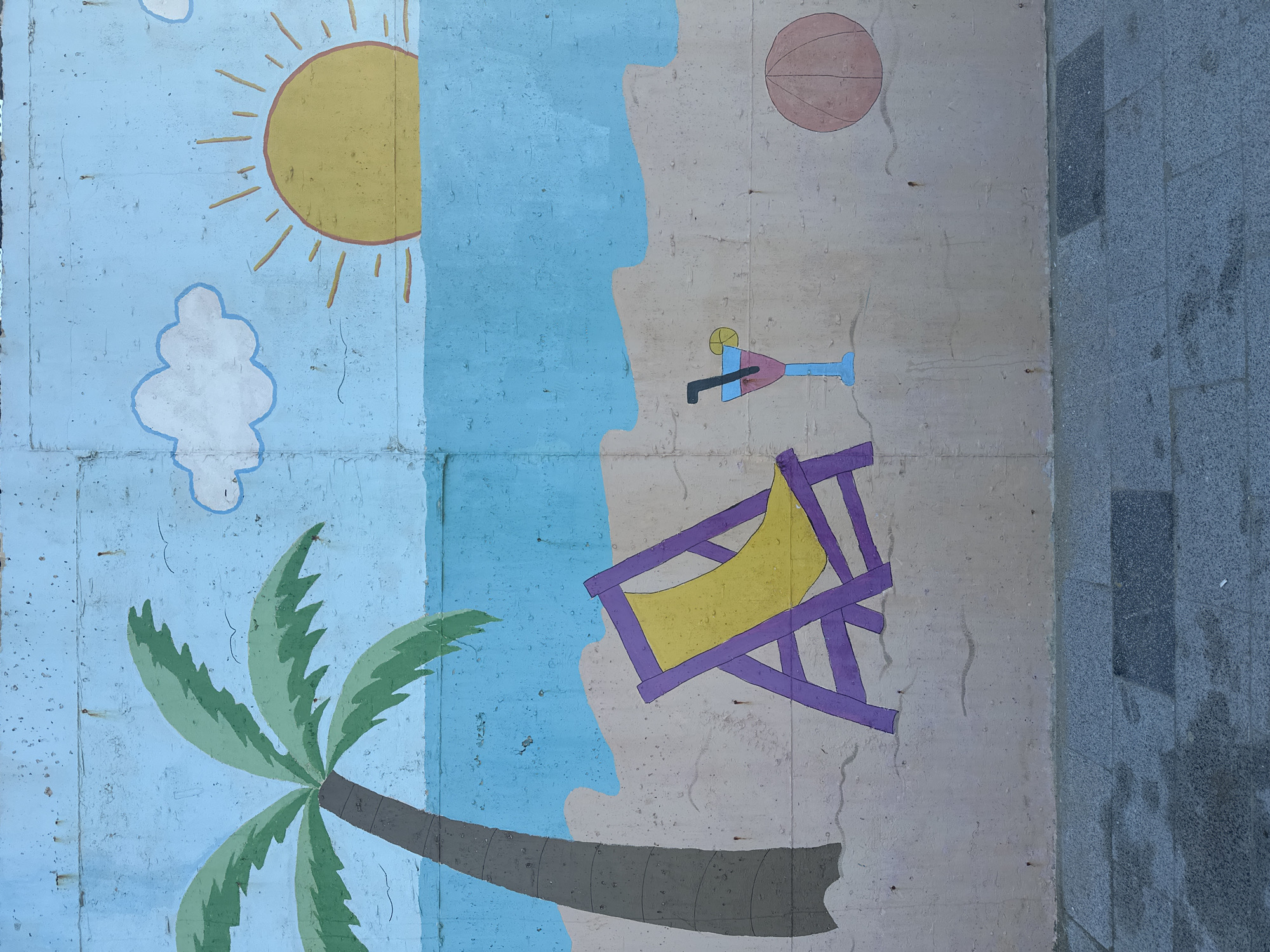 A mural on a concrete wall depicts a beach scene. The left side of the mural shows a blue sky with a yellow sun and a white cloud, and a green palm tree with a brown trunk. A light blue area represents the ocean. The right side represents a sandy beach, including a pink cocktail glass with a blue straw, a beach chair with a yellow seat and purple frame, and an orange beach ball. Gray pavement is visible along the right edge of the image. The location is in the city of Arnavutköy, Turkey.