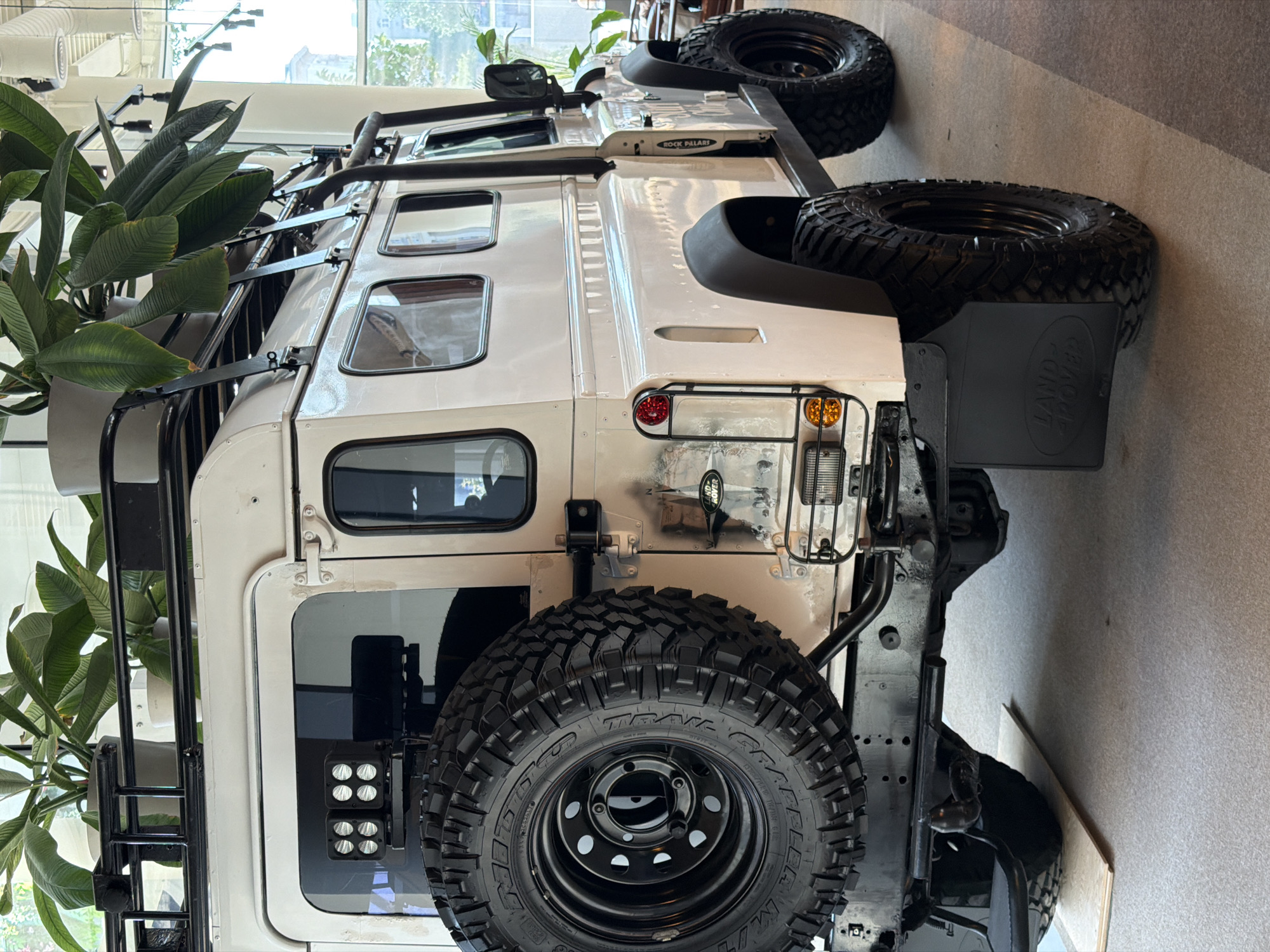 The image shows the back of a light-colored Land Rover Defender with off-road modifications, parked indoors. A large spare tire is mounted on the back. A black ladder is attached to the side of the vehicle. The vehicle has a roof rack and off-road tires. The rear of the vehicle has a Land Rover logo mud flap. The location appears to be indoors with indoor lighting and a plant to the left of the vehicle. There is no indication of the location being in Moscow, Russia from the image.