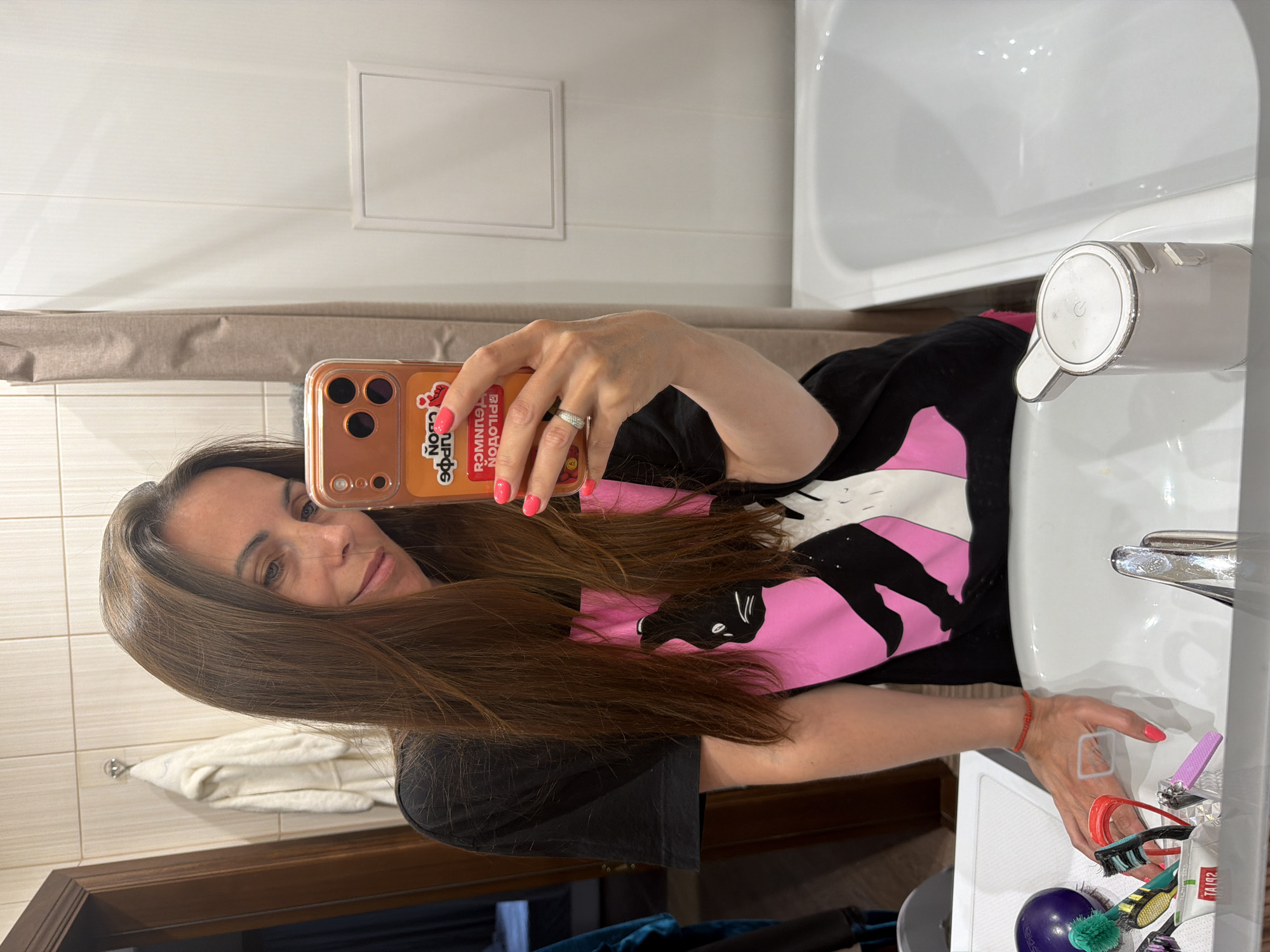 A woman with long brown hair is taking a selfie in a bathroom. She is wearing a black shirt with a pink and black design. Her phone is in a peach-colored case. Her nails are painted pink. There are toiletries and other items in the sink. The location is likely a bathroom in Birobidzhan, Russia.