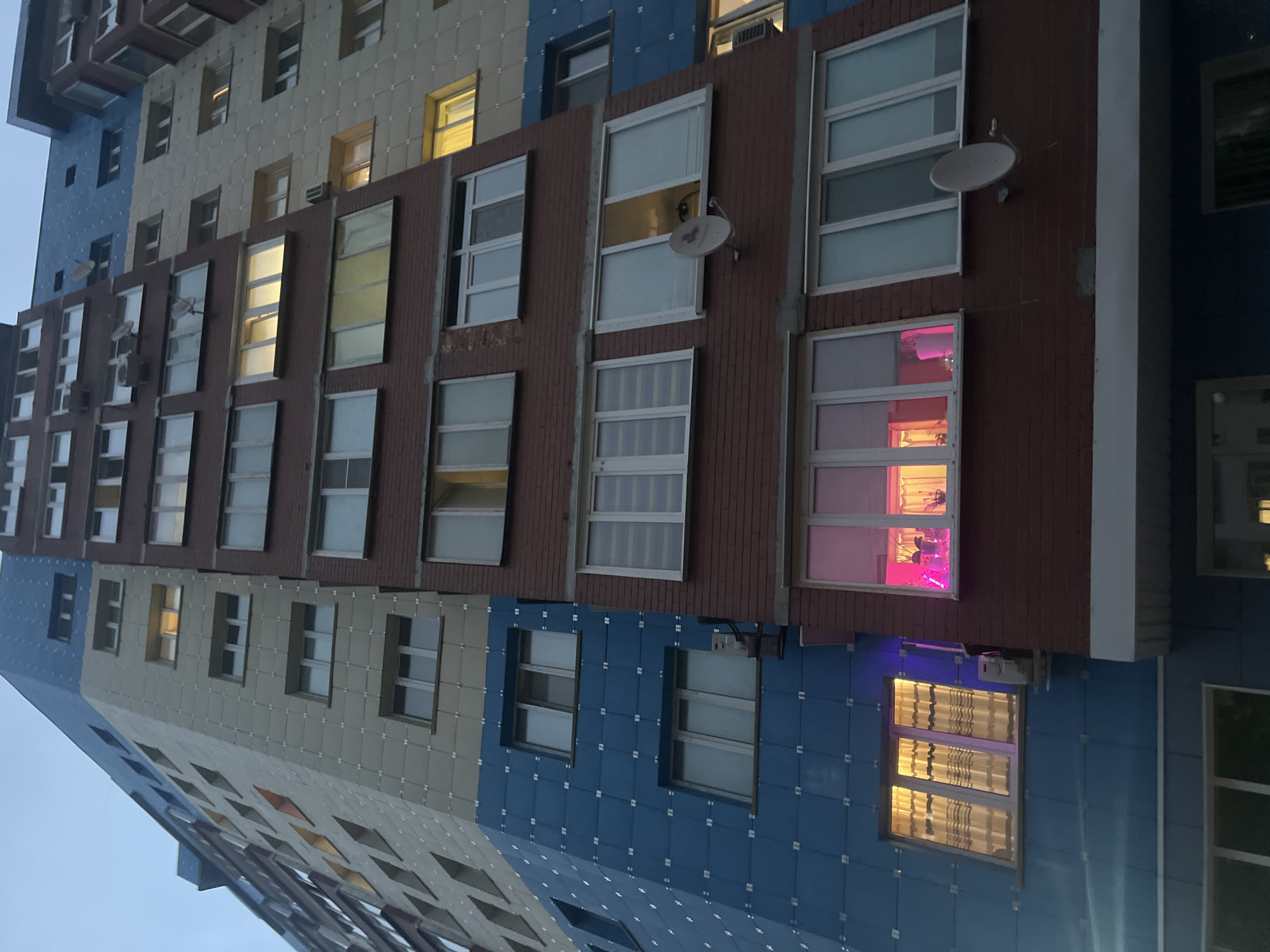 The image shows a multi-story apartment building in Surgut, Russia, with a blue, beige, and red brick facade. The building has numerous windows, some of which are illuminated from within. One window on the blue section of the building has warm yellow light illuminating the interior, with striped curtains visible. Below this, on the red brick section, a window is lit with pink light, revealing a potted plant. Another window on the red brick section shows a satellite dish mounted on the exterior. The sky above is a muted, twilight blue, suggesting the photo was taken around dusk or dawn. The overall impression is of a residential urban environment.