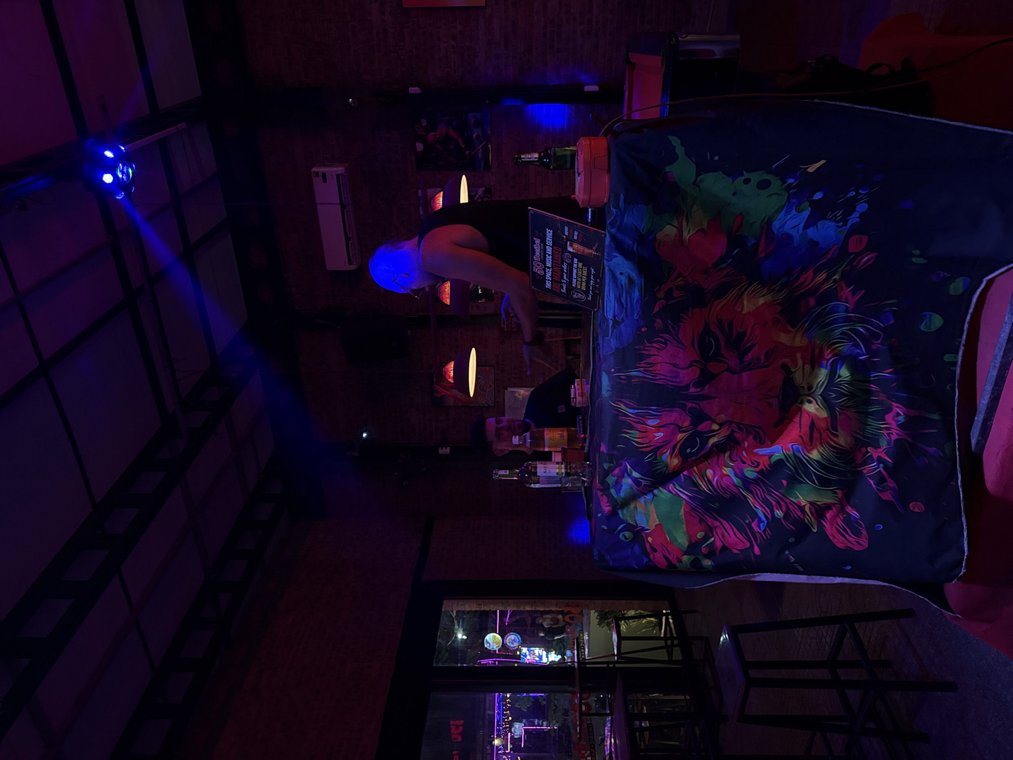 This media file captures the interior of a dimly lit bar or club at night, likely in Đà Nẵng, Vietnam. The scene is dominated by vibrant blue and purple atmospheric lighting.
**Subjects:** A person with short hair, possibly wearing glasses, stands behind a bar or DJ booth, their head illuminated by a blue light. Another individual's head is faintly visible further behind the counter.
**Objects:** The central object is a bar or DJ booth draped with a striking, colorful fabric cover. This cover features an abstract, almost psychedelic design in hues of red, blue, green, yellow, and pink, possibly depicting an animal head like a lion. On the counter, several bottles of spirits, including whiskey, are visible, alongside a white sign. A prominent blue spotlight fixture on the ceiling emits a strong beam of light across the room. An air conditioning unit is mounted on a brick wall, which is adorned with posters or artwork. In the foreground and background, various tables and chairs suggest seating for patrons. A large window or door reveals an urban street scene outside, bustling with distant lights and other establishments, confirming the nighttime setting.
**Activity or event taking place:** The person behind the counter appears to be working, suggesting they are a bartender preparing drinks or a DJ managing music, given the typical setup of such venues. The overall ambiance indicates a social gathering where people enjoy drinks and music.
**Notable details:** The time of day is clearly night, emphasized by the dark interior and the illuminated street. The blue and purple lighting creates a cool, dynamic, and lively atmosphere typical of nightlife. The colorful fabric on the counter adds a unique artistic element to the venue's decor.
**Visible text:** A sign on the counter contains several lines of text, including "50 @centrai", "THIS SPACE, MUSIC AND SERVICE ARE MAINTAINED", "Thanks to your whers", "PLEASE SUPPORT THE BAR", "TONE", and "DRIAK PER GUEST".