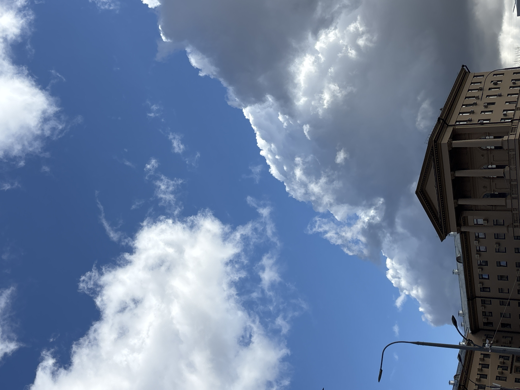 The image is a low-angle shot of a building and the sky. The right side of the image is dominated by the side of a tall building, likely a historic structure with classical architectural elements such as columns and a pediment. The building has multiple windows and air conditioning units visible on its facade. A street lamp extends into the scene from the bottom right corner.
The sky is a bright blue with scattered white clouds on the left side of the frame. On the right side, adjacent to the building, a large, dark grey cloud mass is visible, indicating possible stormy weather.
Without additional information, it is difficult to pinpoint the exact location in Moscow, Russia.