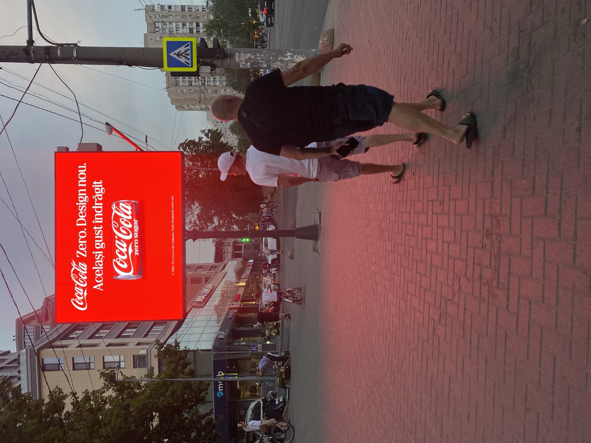 The image depicts an outdoor scene in Chișinău, Moldova, possibly on a street or pedestrian walkway. A large digital billboard displays a Coca-Cola Zero advertisement with the text "Coca-Cola Zero. Design nou. Același gust îndrăgit." (New design. Same beloved taste.) and an image of a Coca-Cola can. Below the main advertisement, there is smaller text stating "2020, The Coca-Cola Company. Toate drepturile rezervate." (2020, The Coca-Cola Company. All rights reserved.).
In the foreground, two men are walking away from the camera on a brick-patterned sidewalk illuminated by a reddish hue, likely from the billboard's light. The man on the left wears a white t-shirt, grey cargo shorts, and flip-flops. The man on the right wears a black t-shirt, dark shorts, and flip-flops.
In the background, there are several buildings, including a modern building with glass facades and a taller apartment building. A street with cars and a pedestrian crossing with a traffic signal and a pedestrian crossing sign are visible. Other pedestrians and cyclists are also present in the mid-ground and background, suggesting a busy urban environment. The sky is overcast, indicating either early morning or late evening.
