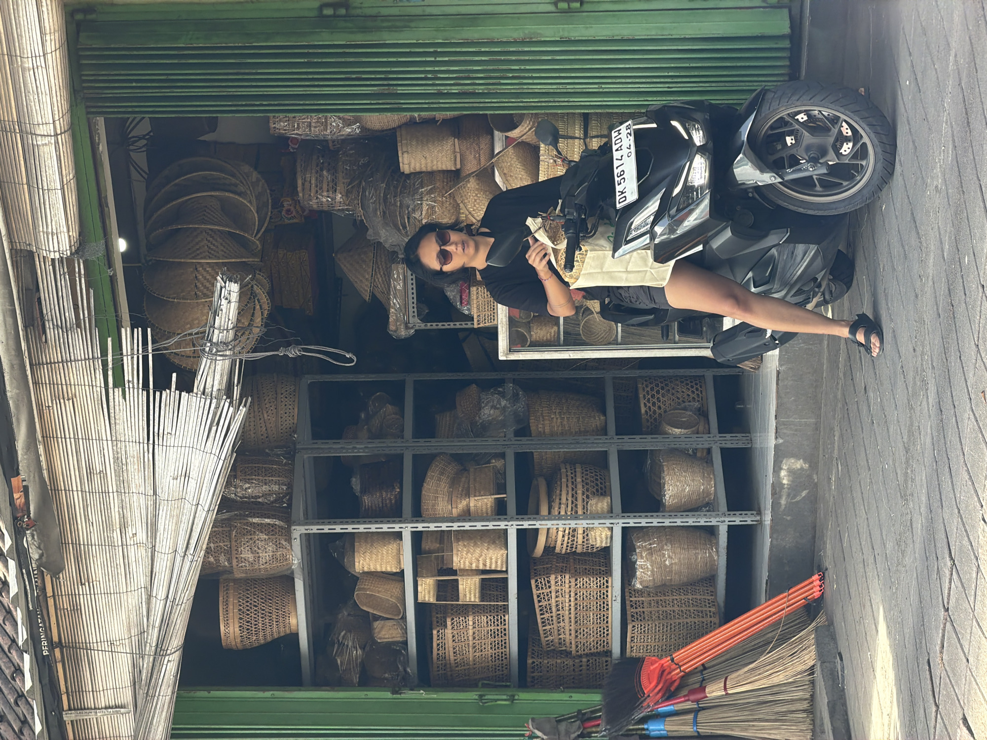 The photo depicts a woman sitting on a parked motorcycle in front of a store in North Kuta, Indonesia. The store is filled with baskets of various shapes and sizes, some stacked on shelves and others hanging from the ceiling. The woman wears a black shirt, sunglasses, and appears to be holding something in her hand. The motorcycle is black and gray with a license plate visible, but not entirely legible. The storefront has a green metal shutter partially open. Brooms rest on the lower right corner.