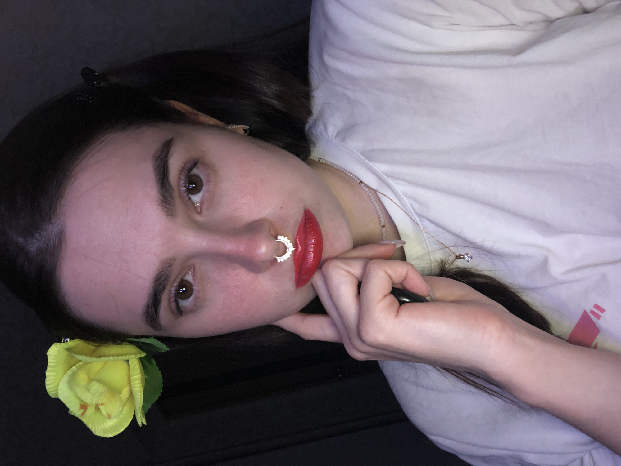 A young woman with brown hair and fair skin is lying down, her head tilted to the left, resting on her hand. She is wearing a white t-shirt, a beaded necklace, and a gold pendant necklace. She has brown eyes, dark eyebrows, and is wearing bright red lipstick. She has a silver septum piercing and a yellow flower pinned in her hair near her left ear. She is holding a black object in her hand. The background is dark. There is no indication this photo was taken in the city of Noviy, Russia.