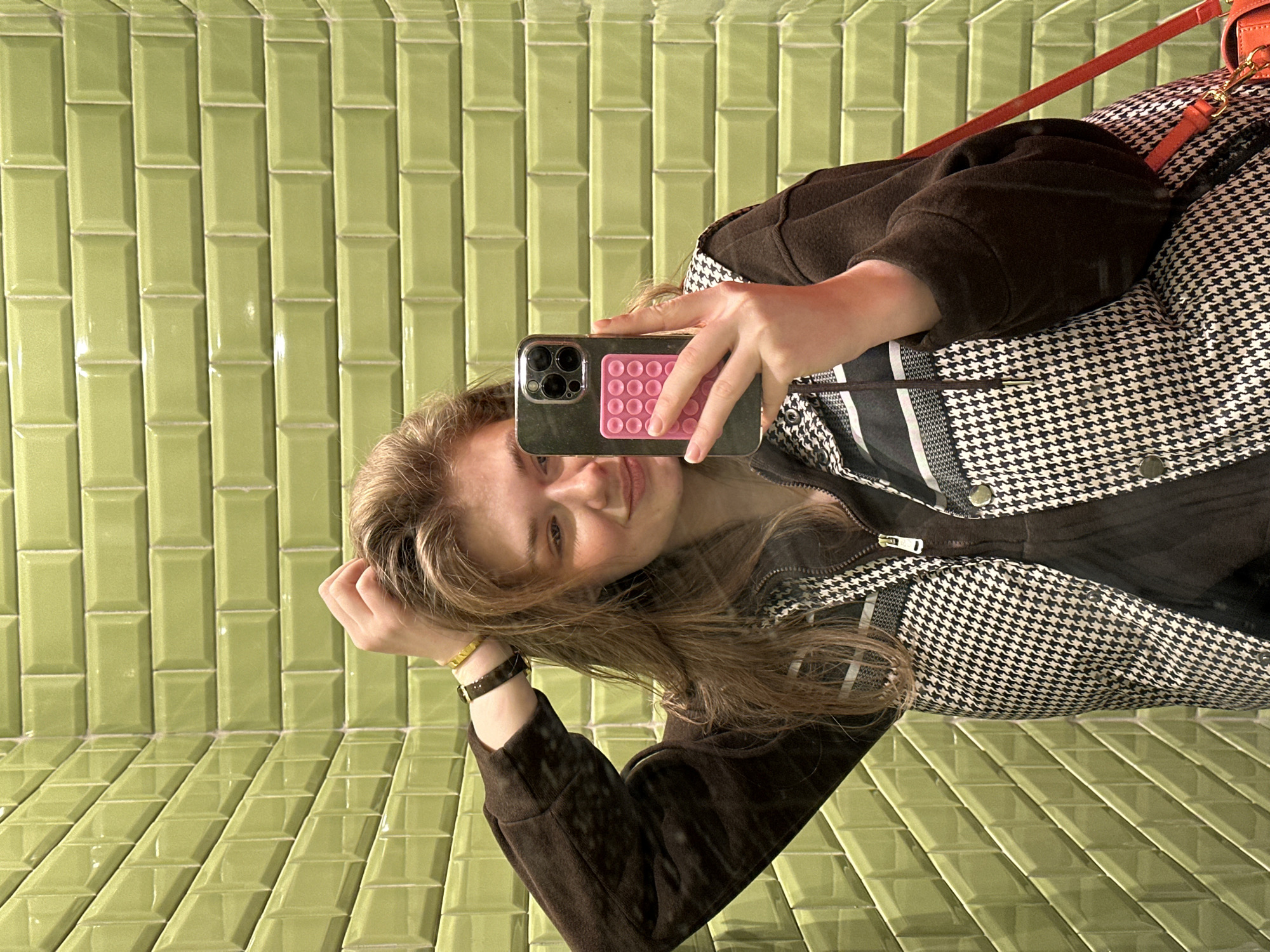 A young adult female is depicted in a mirror selfie, smiling subtly, within an interior space located in Kazan, Russia. She is shown from the torso up, holding a dark-cased smartphone, equipped with a pink, multi-button sensory accessory on its back, in her right hand, pointed towards the mirror to capture the image. Her left hand is positioned with fingers near her temple, lightly touching her light brown hair. She wears a dark brown long-sleeved top beneath a black-and-white houndstooth patterned gilet featuring a central zipper and horizontal stripe details on the upper chest. A gold-colored bracelet and a dark wristband are visible on her left wrist. A segment of an orange-red shoulder bag strap is visible on her right side. The background environment consists of two walls covered entirely in glossy, light green subway tiles. The tiles on the wall behind her right side are arranged horizontally, while those on the wall behind her left side are arranged diagonally, meeting at a distinct corner with white grout lines.