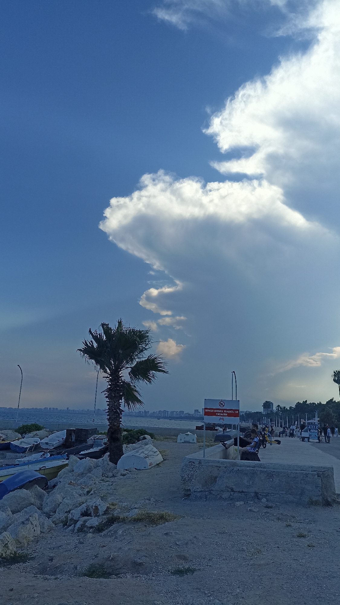The image depicts a coastal scene in Yenişehir, Turkey, under a vast blue sky with dramatic, towering white clouds. In the foreground, a sandy and rocky ground leads to the edge of the water, where several boats are pulled ashore. These boats, of various sizes and colors, are partially covered and rest on a bed of white rocks and some scattered debris.
A lone palm tree with full fronds stands prominently on the left side of the frame, its trunk dark against the lighter ground. To the right of the palm tree, a concrete structure with a rough texture forms a low wall or seating area. Behind this, a paved walkway stretches into the distance, lined with more rocks and vegetation.
A sign with red and white lettering is mounted on a pole, indicating a prohibition related to entering the sea. Further along the walkway, several people are visible. Some are seated on benches, while others are standing or walking, some interacting with each other. There are also what appear to be bicycles and a small cart on the walkway. In the background, a city skyline is visible across the water, with buildings extending along the coast. The sea itself is calm, with a faint horizon line separating it from the distant land. The overall lighting suggests it is either early morning or late afternoon.