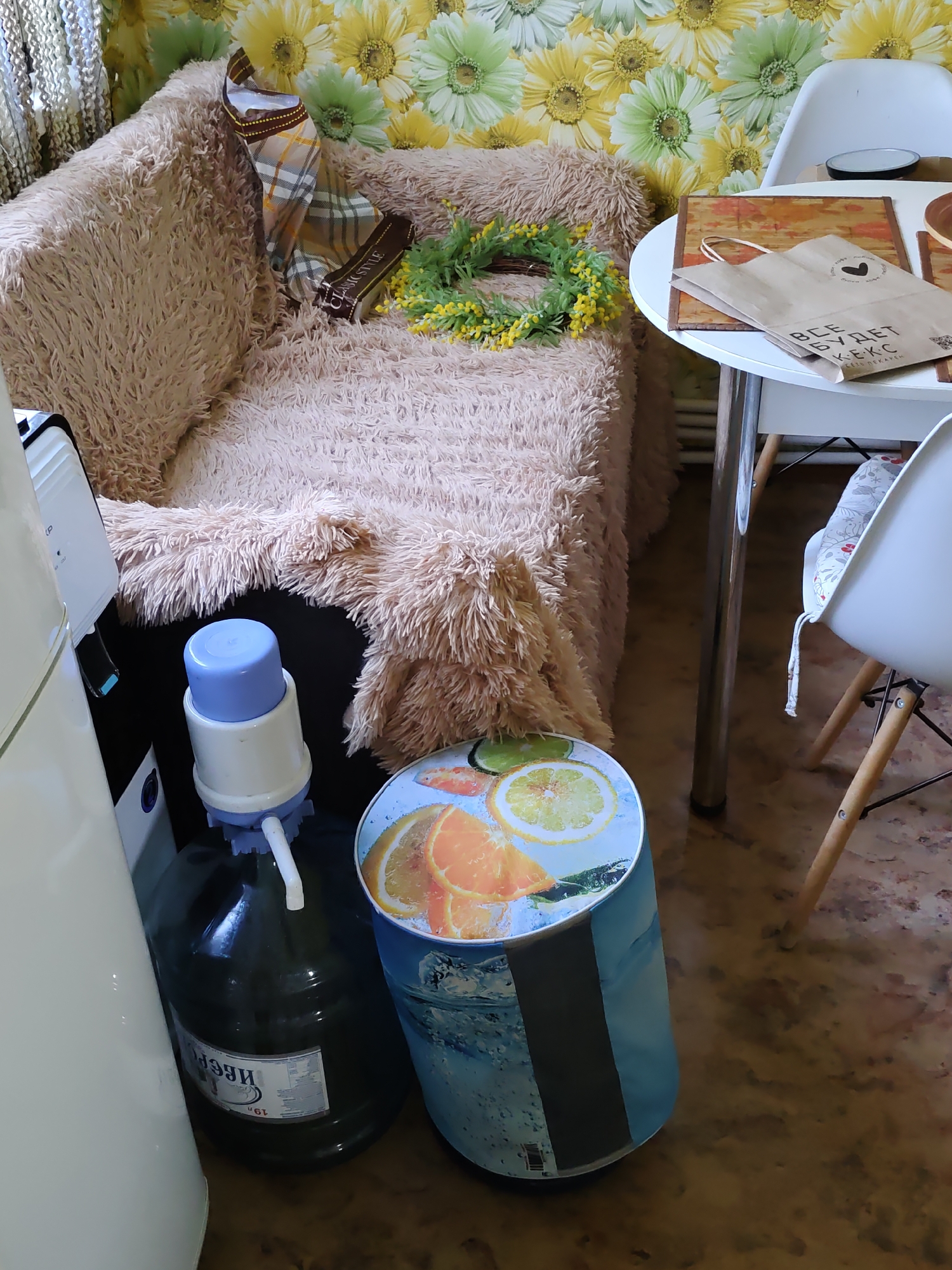 The image captures an indoor scene, likely a domestic setting, with a focus on a plush, beige, faux-fur armchair covered with a plaid scarf and a small yellow wreath. To the right of the armchair is a white round table with a brown placemat, a paper shopping bag with Cyrillic text, and a wooden bowl. The background features wallpaper with a floral pattern of yellow and green flowers. In the foreground, a large dark green plastic bottle with a blue manual water pump sits on the floor next to a cylindrical container decorated with images of citrus fruits and water. A portion of a white appliance, possibly a refrigerator or water cooler, is visible on the far left. The floor appears to be a speckled, reddish-brown linoleum. The image composition suggests a casual, possibly cluttered, living space. No specific landmarks or clues point to a precise location within Rostov-on-Don, Russia.