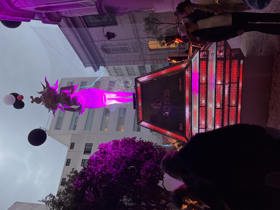 A vibrant scene shows a sculpture of a woman with a radiant purple glow, seemingly floating with a cluster of balloons. Behind the sculpture are buildings, adding depth to the urban setting. To the right, a geometric, pyramid-shaped structure illuminated in red and white displays "SABINO GURSU" and "AYNOS." In the foreground, a bush with purple foliage and silhouettes of people suggest a gathering of observers enjoying the unique installation.